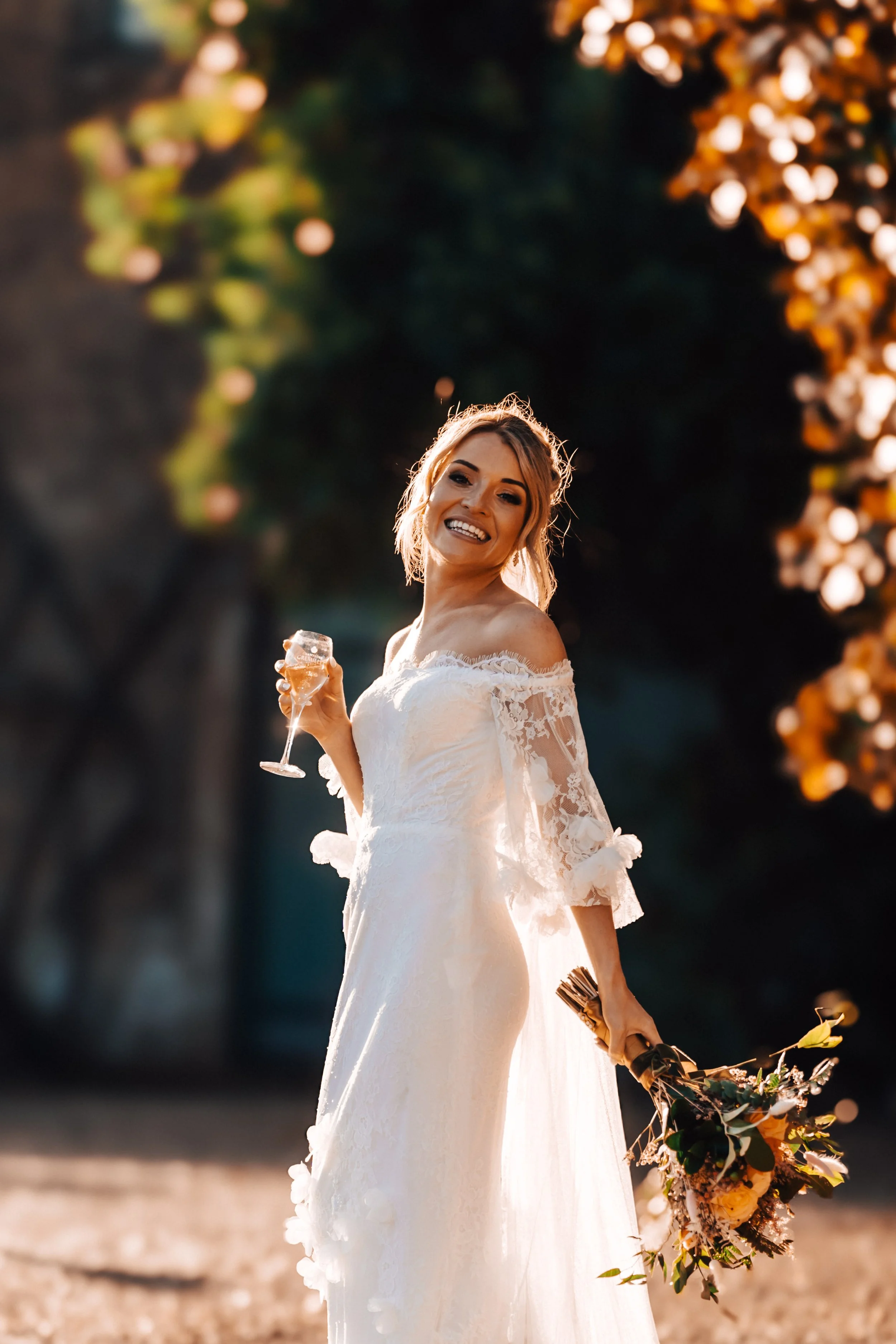 A bride in a white lace wedding dress holding a glass of champagne and a bouquet, smiling outdoors during sunset.