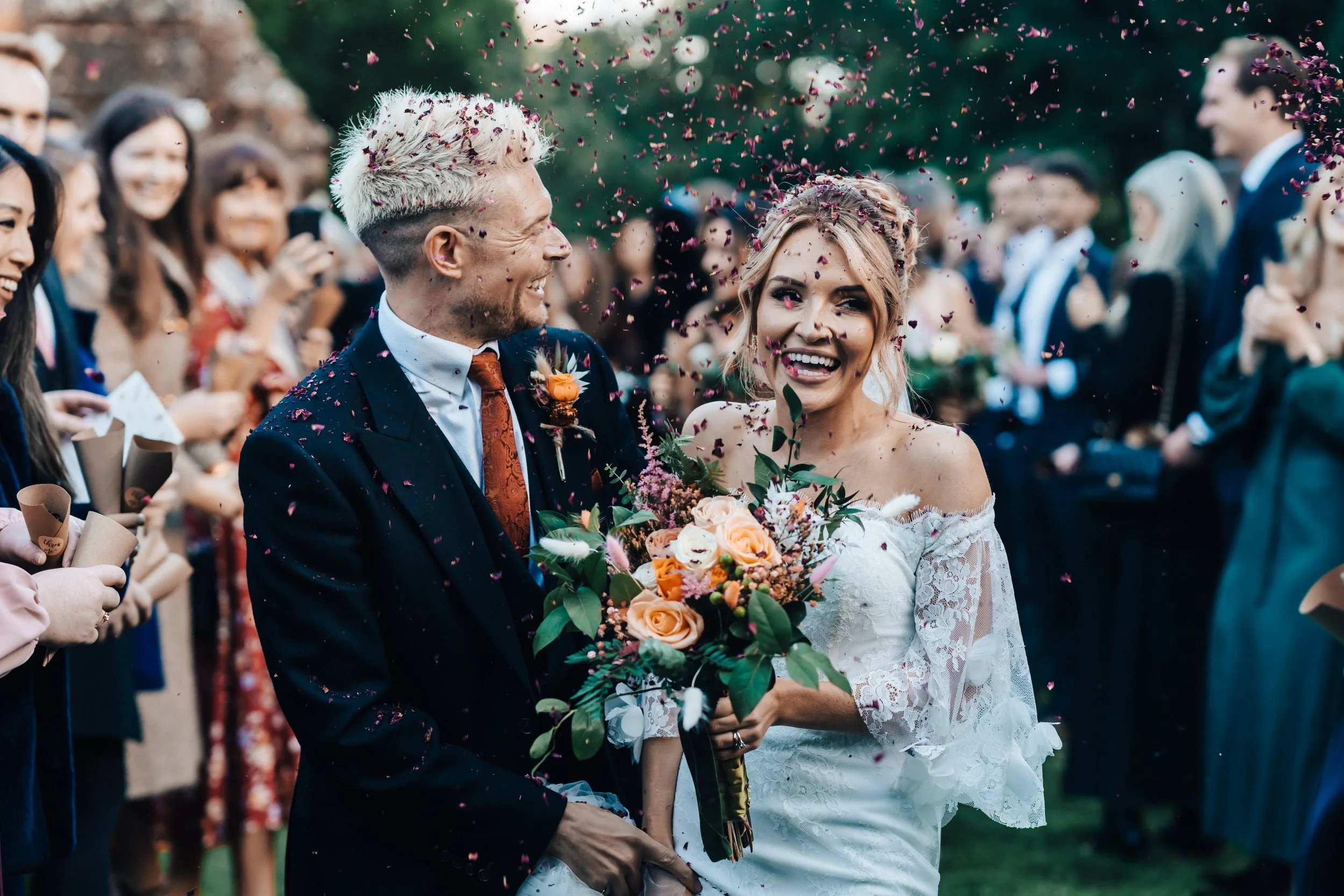 A bride and groom at their wedding ceremony, smiling and holding a bouquet, surrounded by friends and family, with confetti in the air.