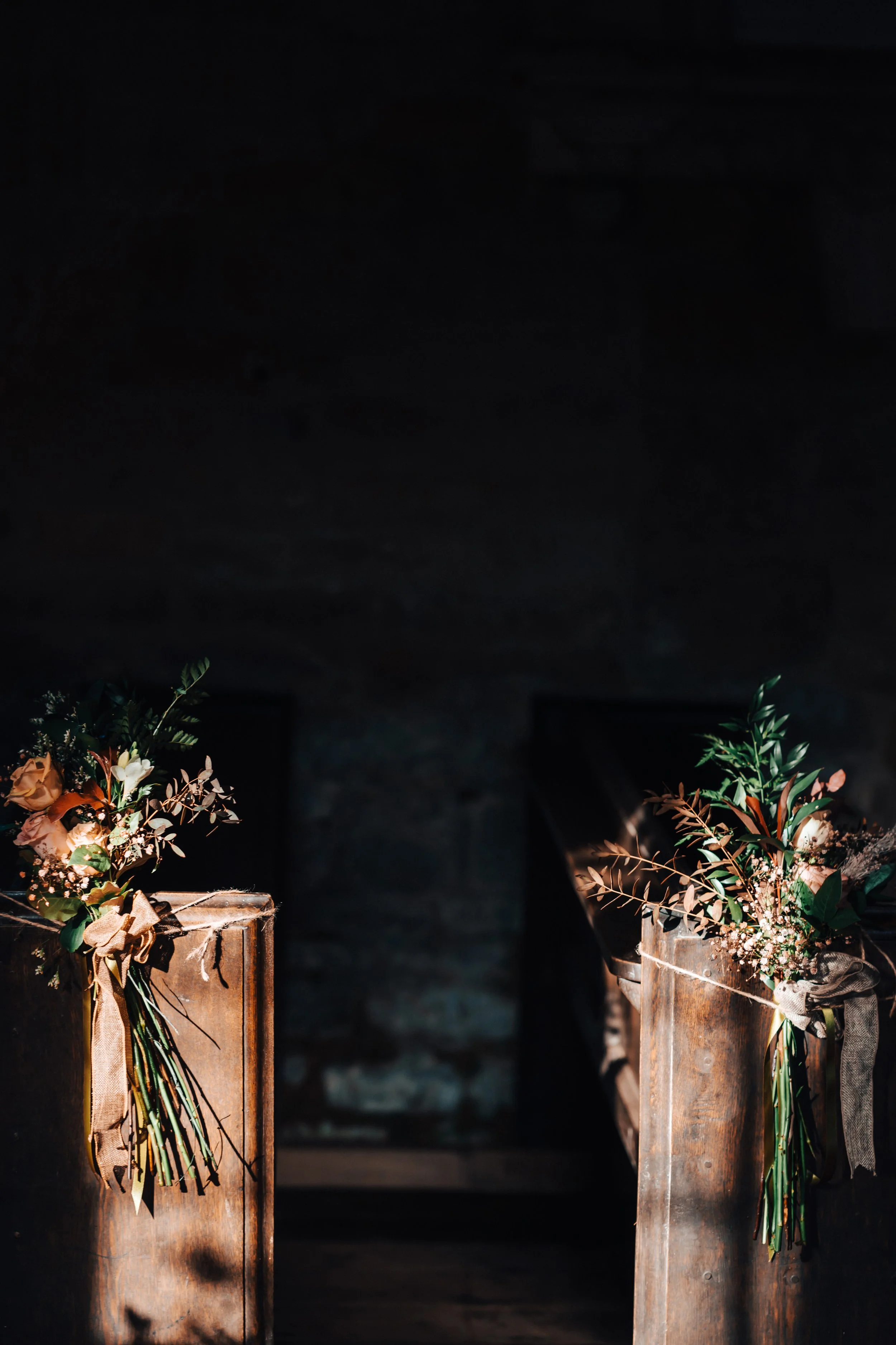 Wooden church pews decorated with flower arrangements of roses, greenery, and small white flowers, tied with burlap and ribbon, inside a dimly lit venue.