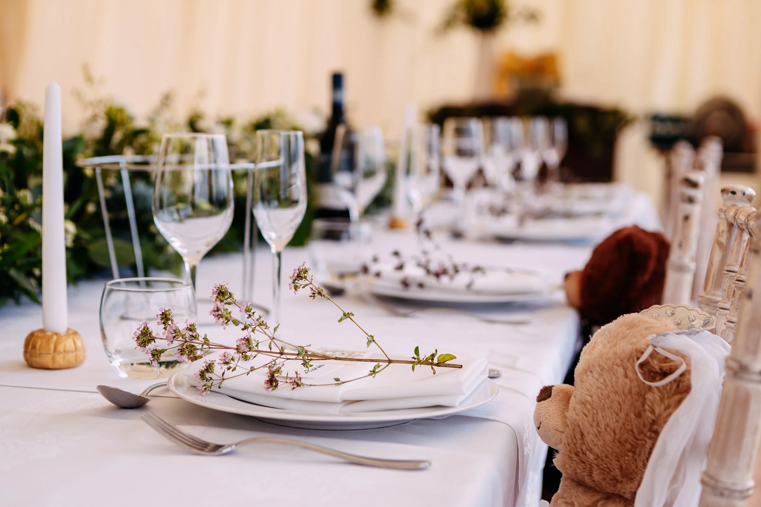 Decorated dining table set for a special occasion with glasses, plates, a white napkin with a sprig of pink flowers, candles, and teddy bears on some chairs, in an elegant indoor setting.