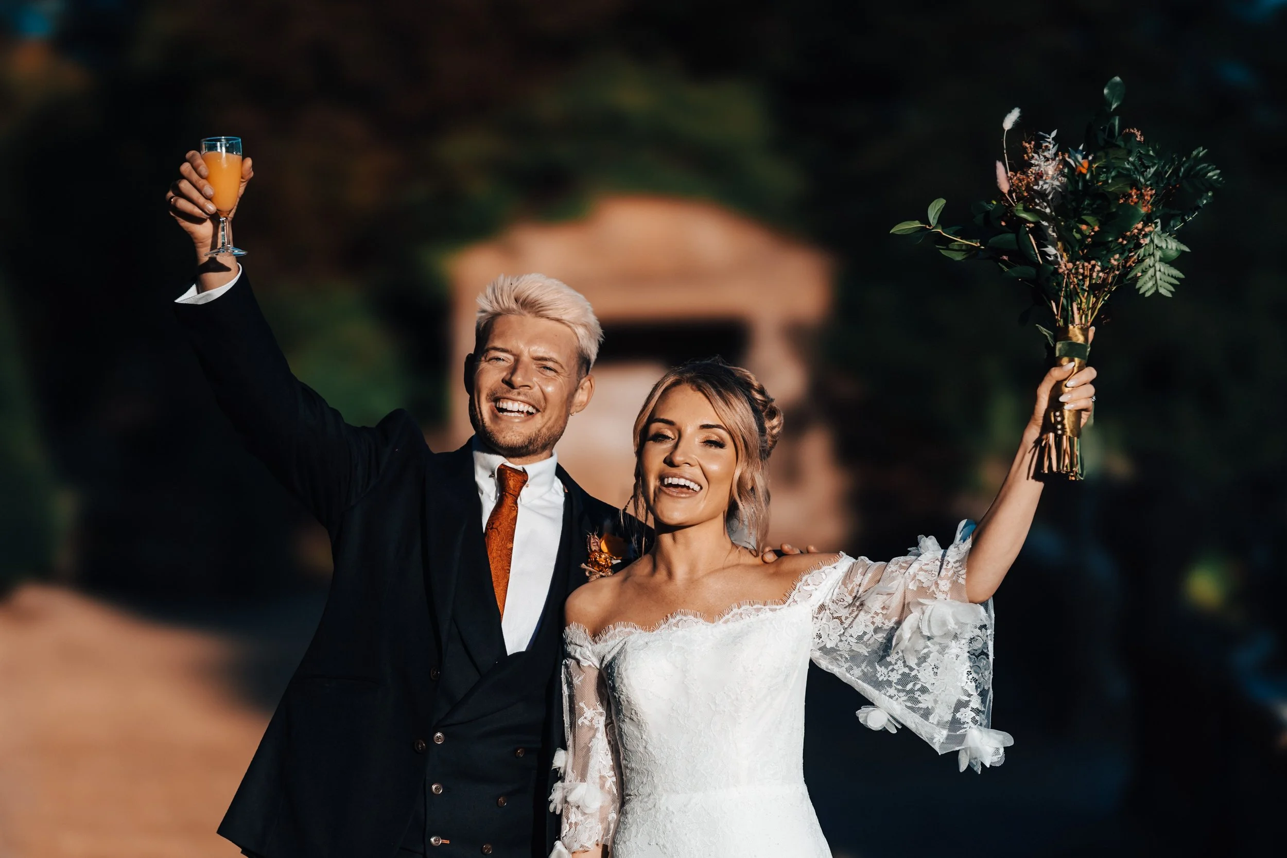 A newlywed couple celebrating outdoors, smiling and holding drinks and a bouquet, with a blurred background.