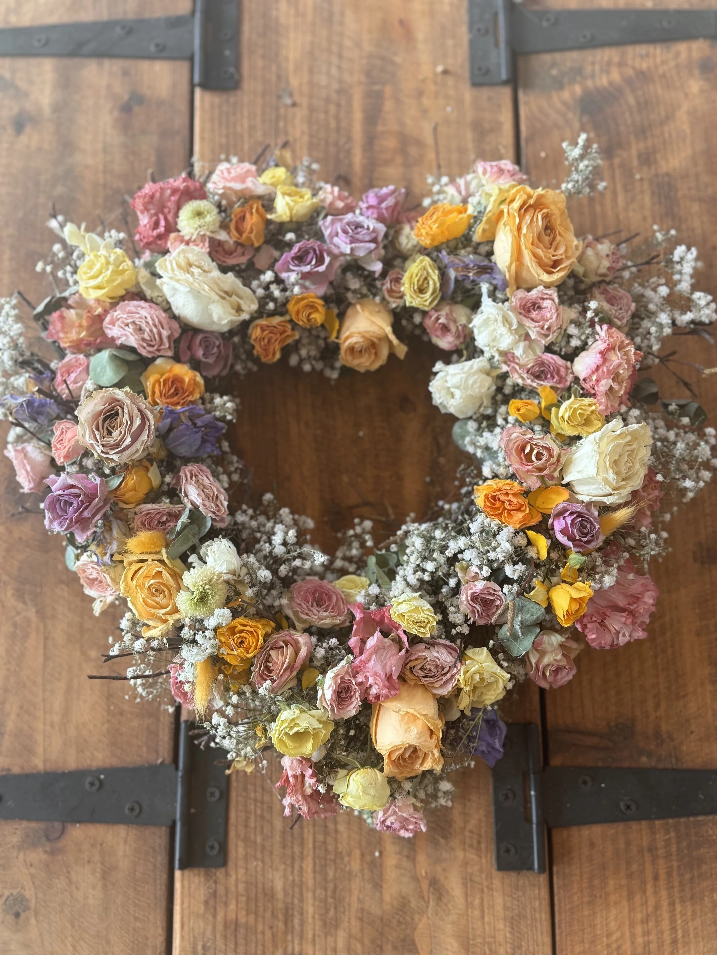 Floral heart-shaped wreath made of pink, yellow, orange, purple, and white roses with baby's breath, on a wooden table.
