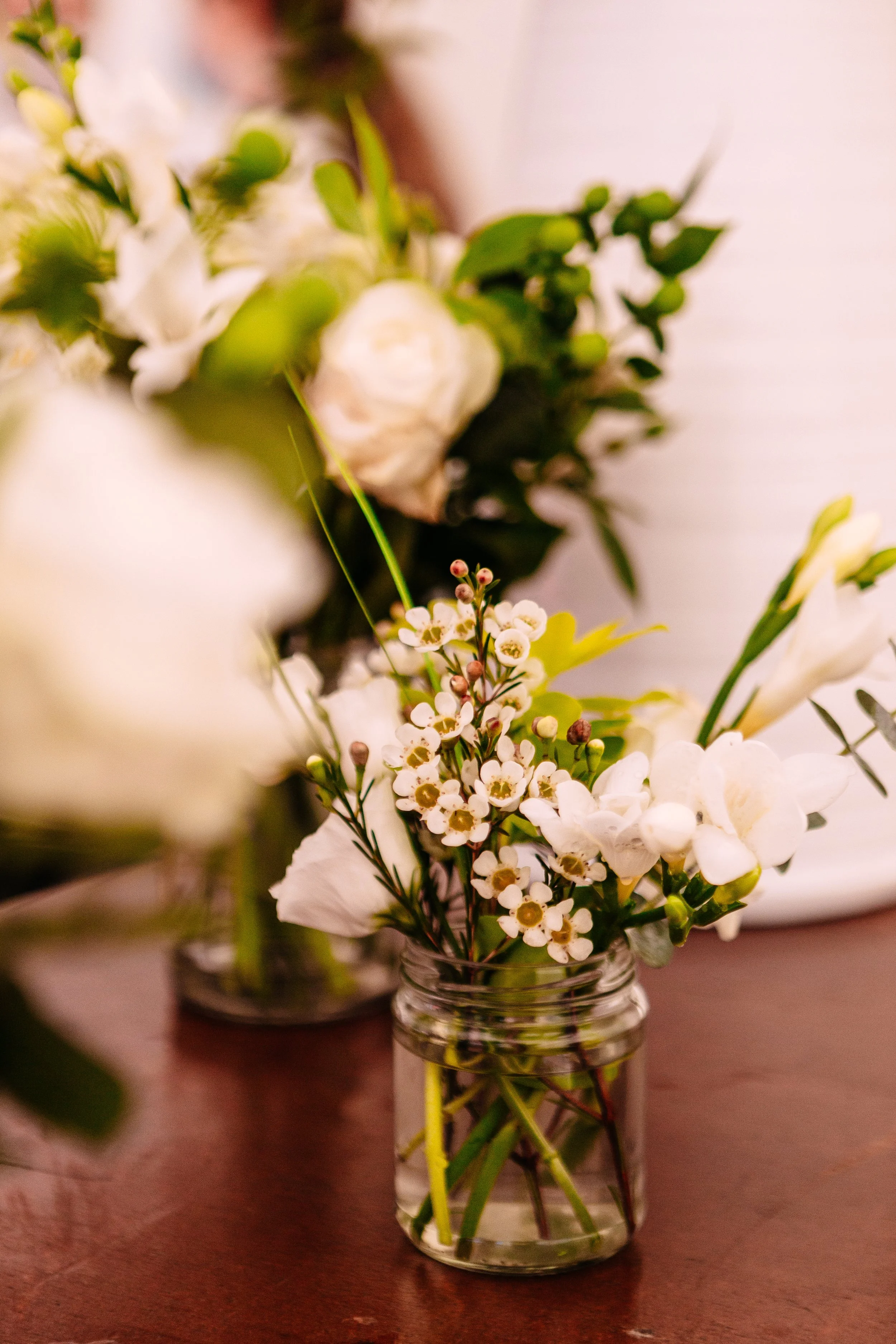 Small bouquet of white flowers in a glass jar on a wooden surface.
