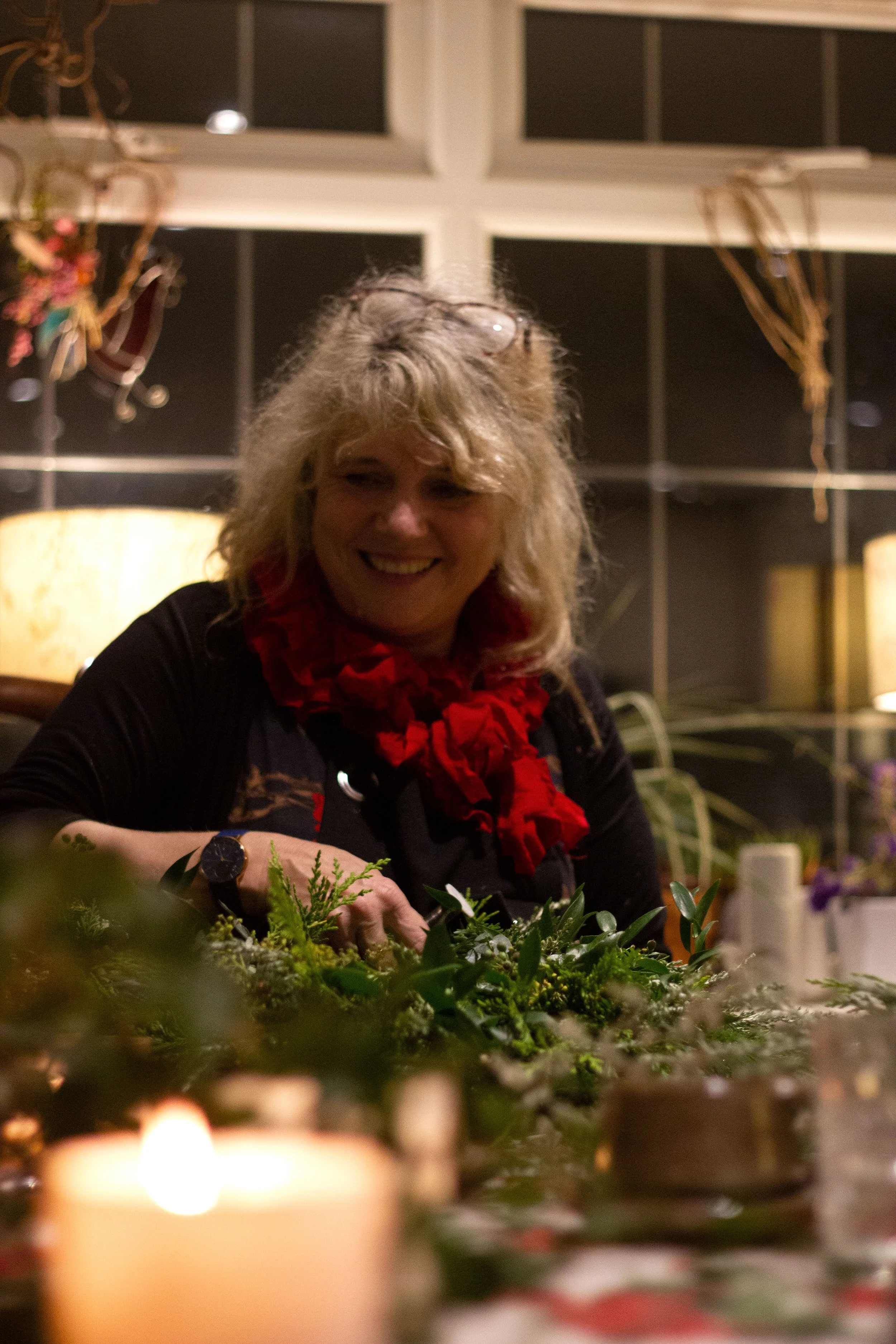 A woman with curly blonde hair, wearing glasses on her head and a red scarf, smiling while arranging greenery on a table, indoors with dark windows and warm lighting.