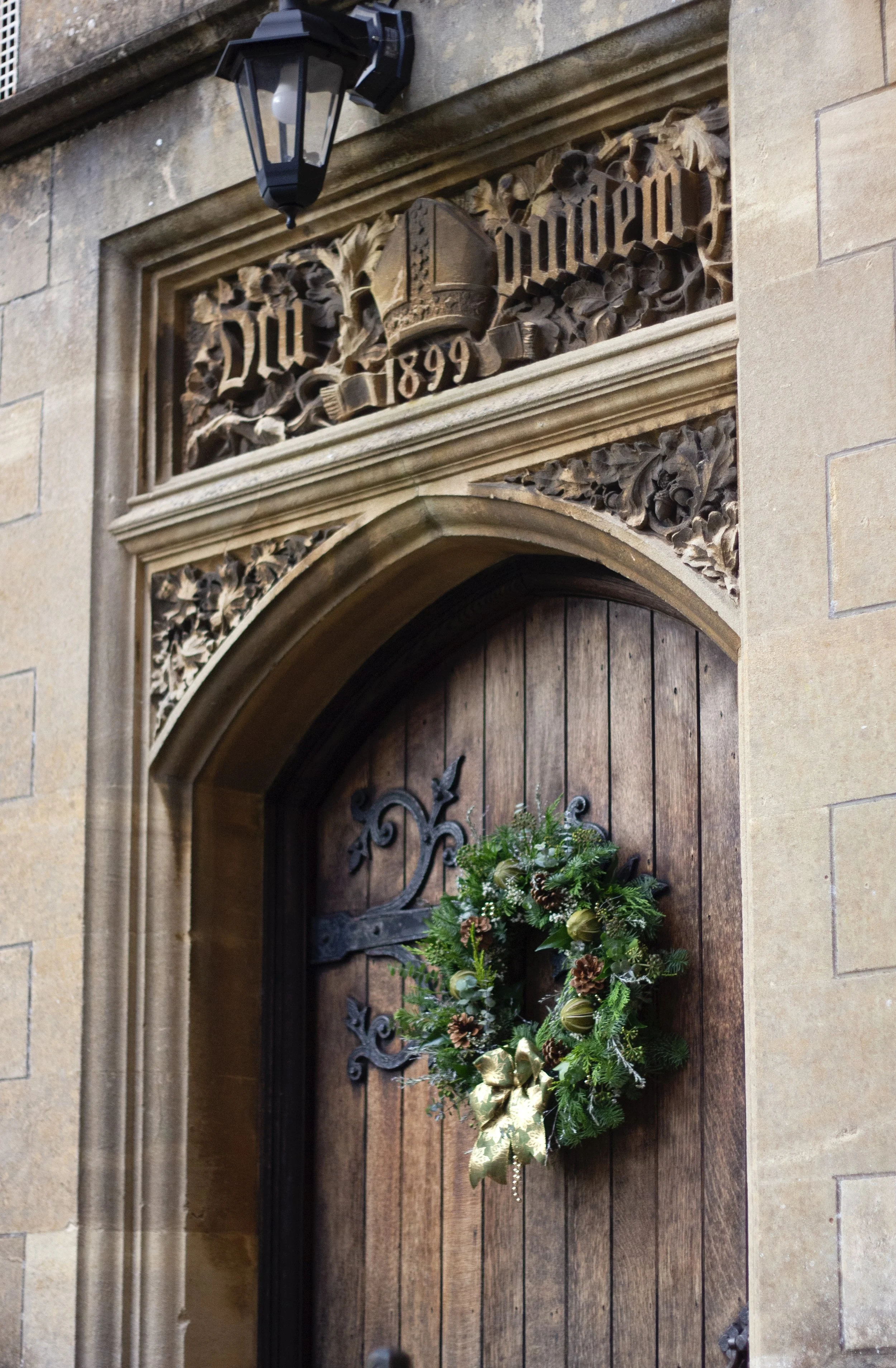 A wooden door decorated with a festive Christmas wreath, surrounded by stone architecture and an ornate carved stone plaque with the year 1899 and a shield emblem.