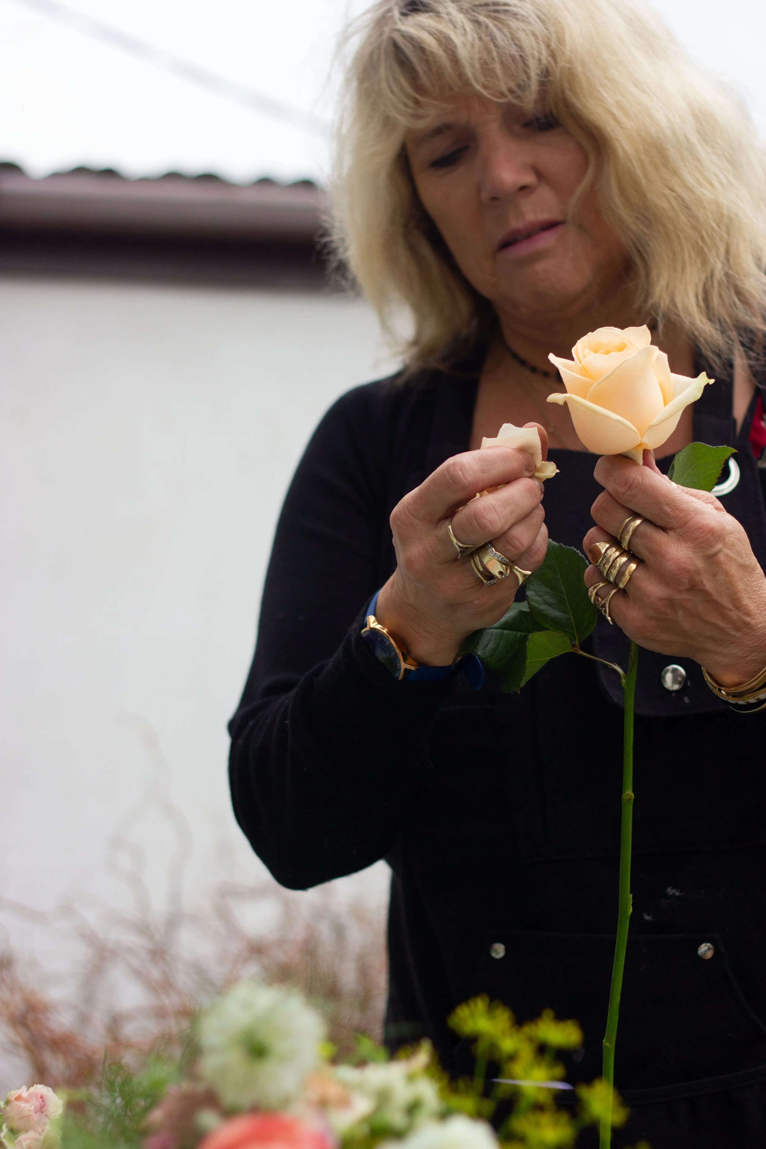 A woman with blonde hair examines a pale peach-colored rose in her hand, wearing rings and a black top, with a blurred outdoor background.