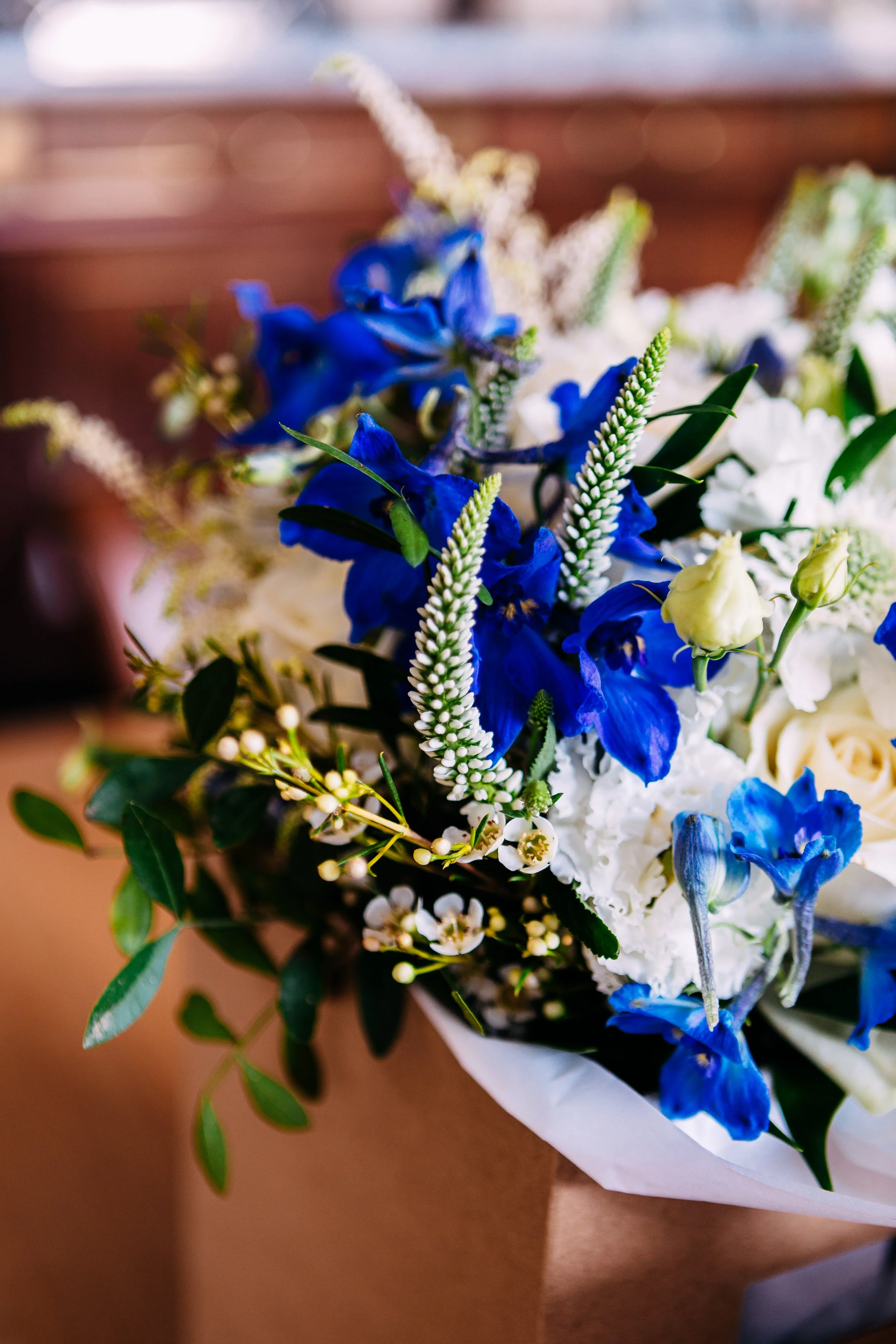 Close-up of a colorful flower arrangement with blue, white, and cream flowers, joined by green leaves, wrapped in brown paper.