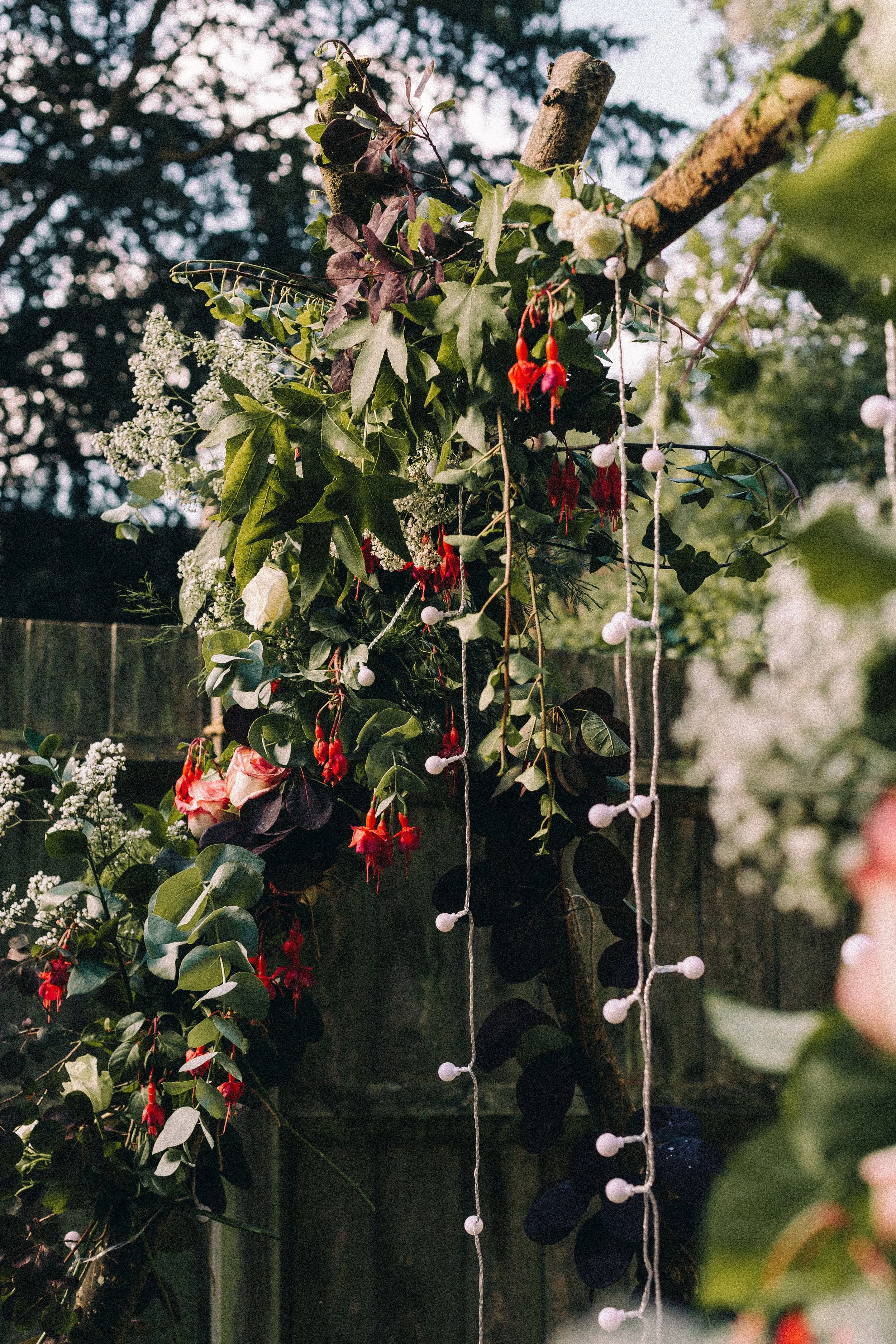 Decorative outdoor floral arrangement with various green, red, and white flowers and leaves, with string lights hanging from a fence.