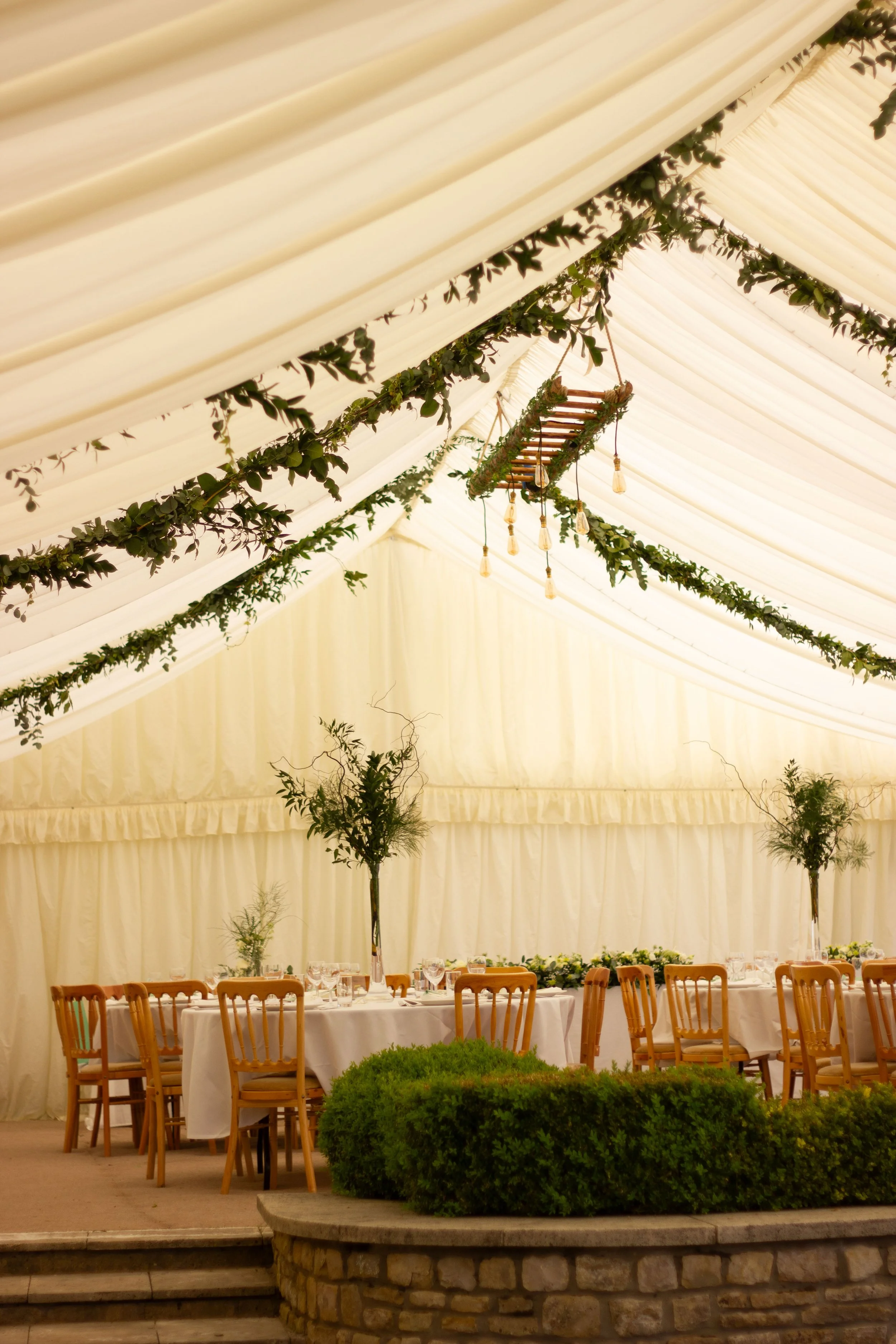 Decorated banquet tent with draped fabric, greenery, hanging lights, tables with white tablecloths, wooden chairs, and potted plants.