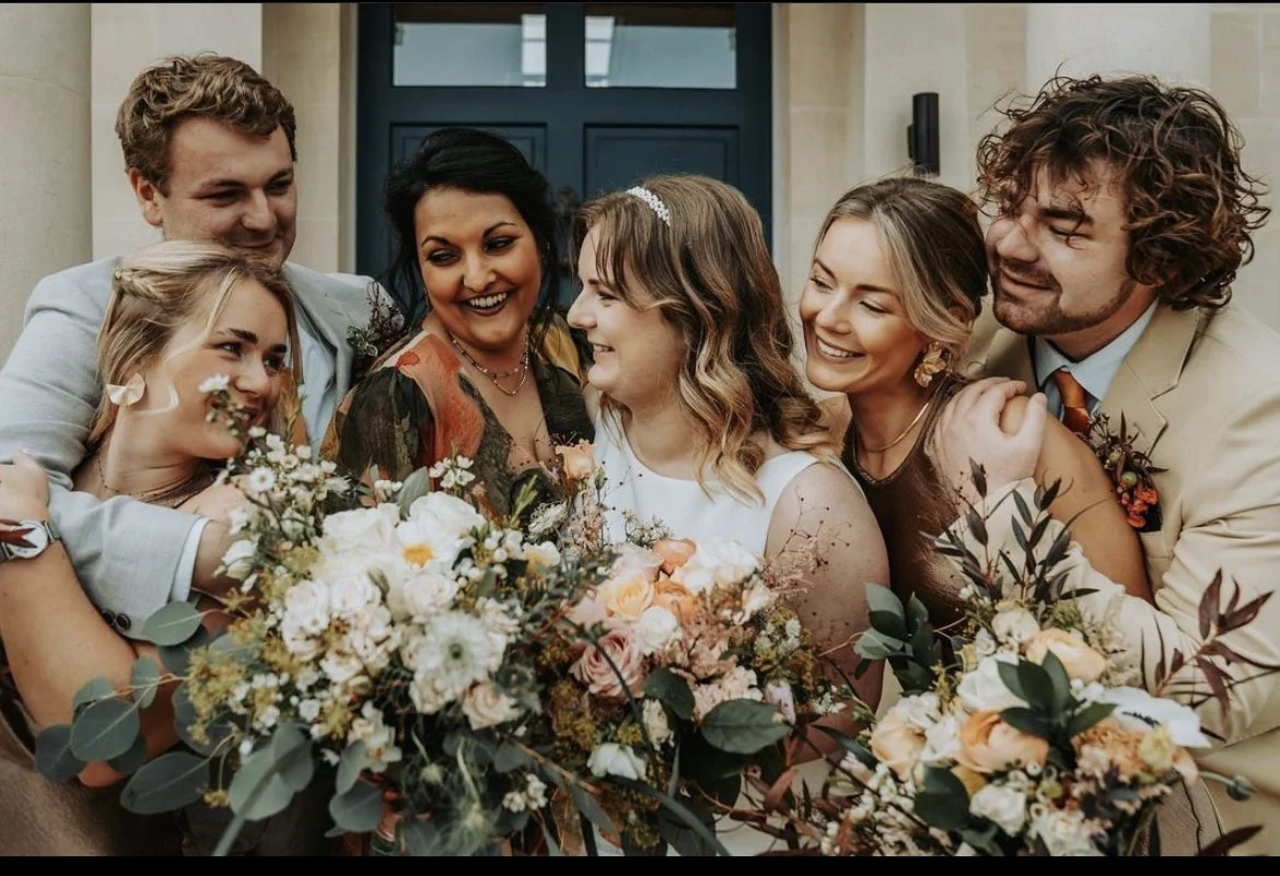 Group of six people celebrating a wedding, smiling and hugging, with a large bouquet of flowers in front, in front of a building with a blue door.
