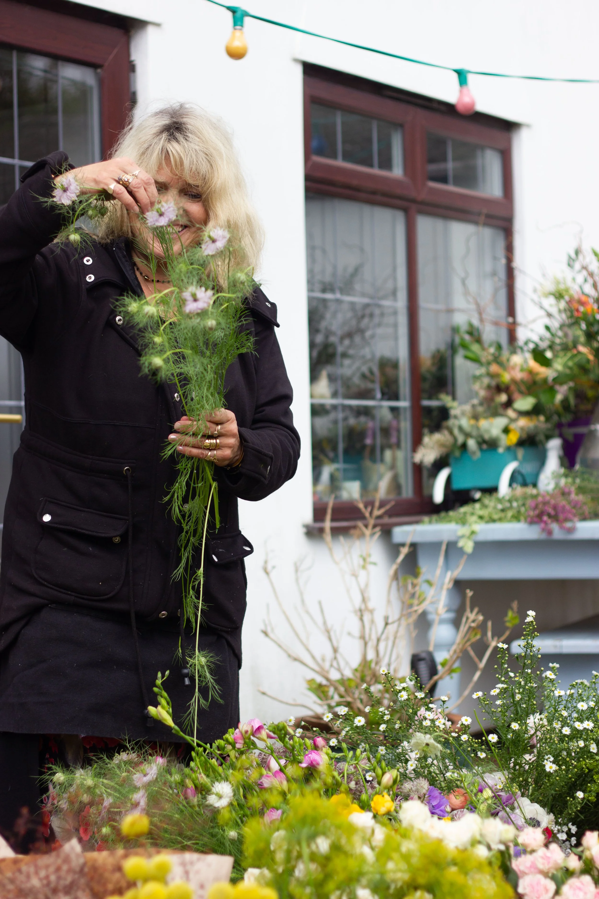 A woman with blonde hair wearing a black jacket is gathering flowers in her garden. There are colorful flowers and plants around her, and a house with large windows and multicolored string lights hanging above.