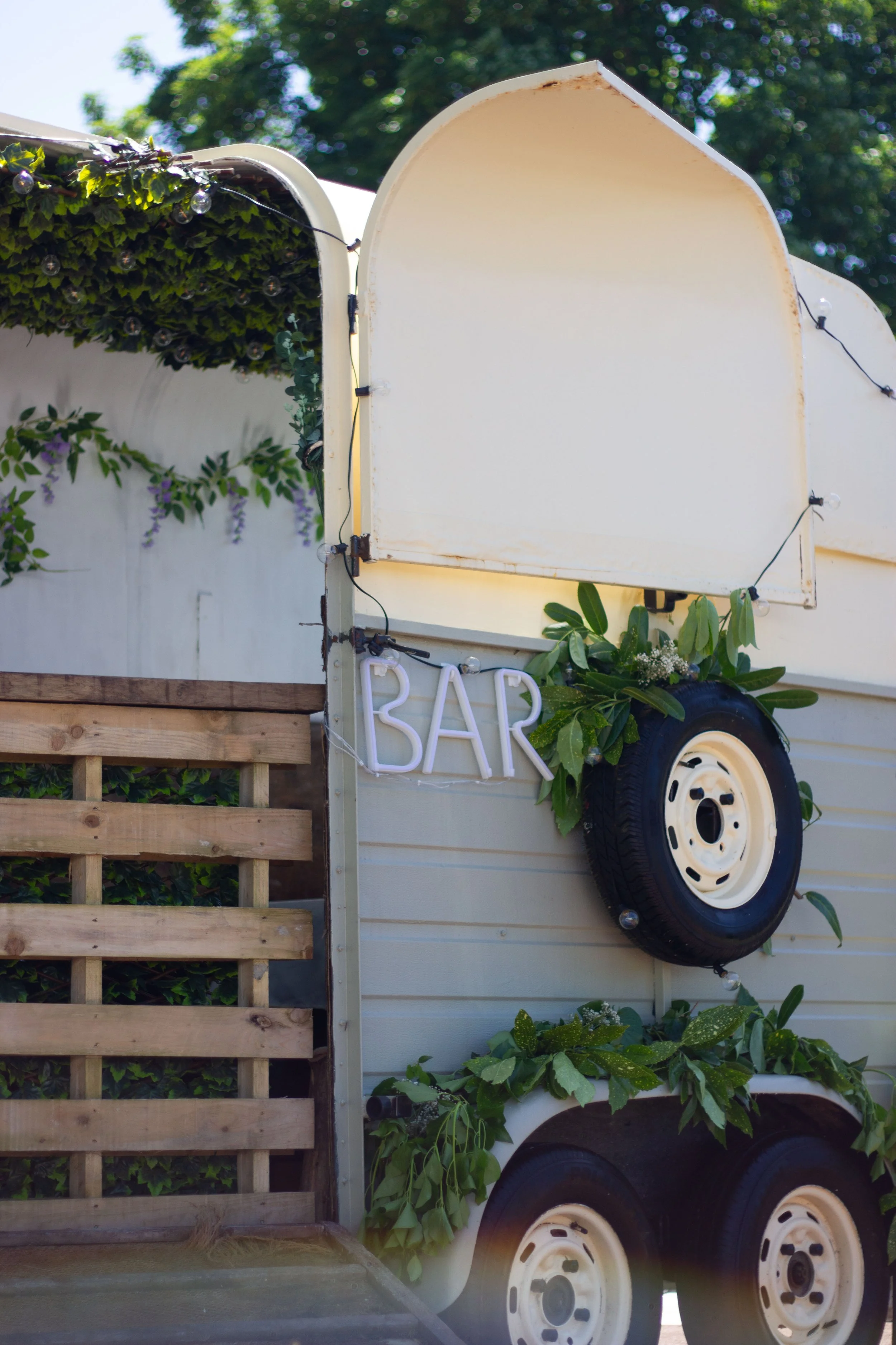 A white mobile bar trailer decorated with greenery, a tire, and a neon sign spelling 'BAR' in capital letters.