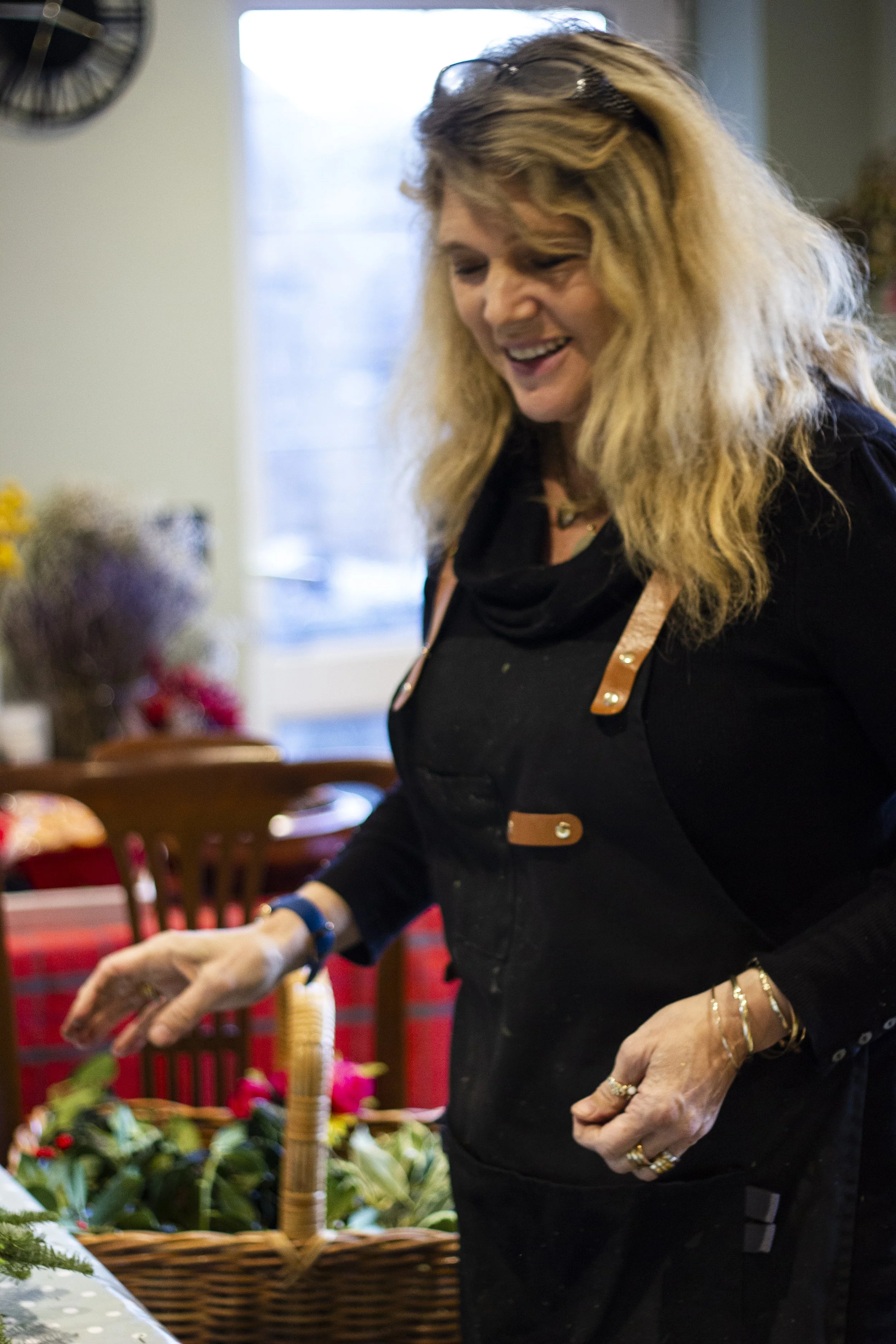 A woman with blonde, wavy hair and glasses on her head smiling while arranging flowers in a wicker basket inside a room with a window and a wall clock.