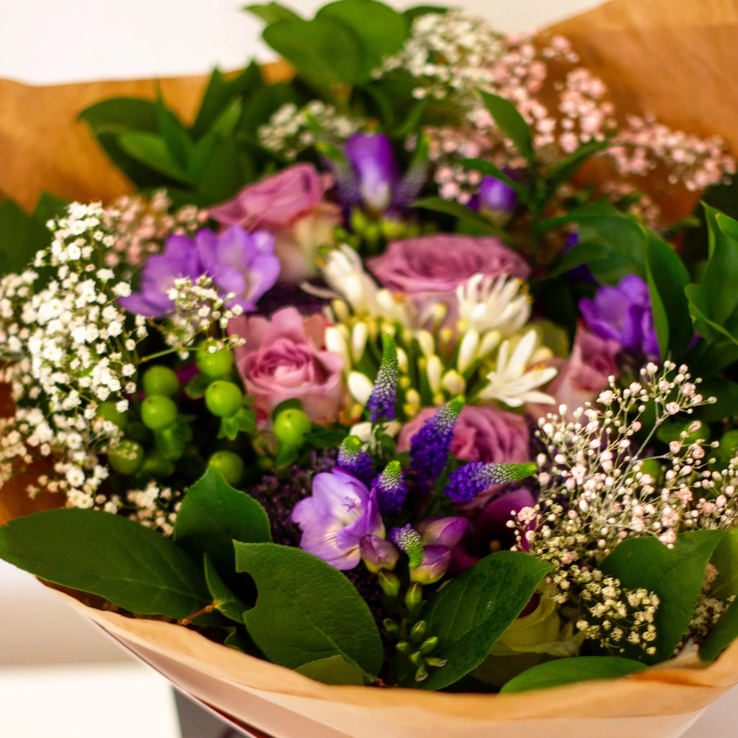 A close-up of a colorful bouquet of flowers including purple, pink, and white blossoms, green leaves, and small white filler flowers wrapped in light brown paper.