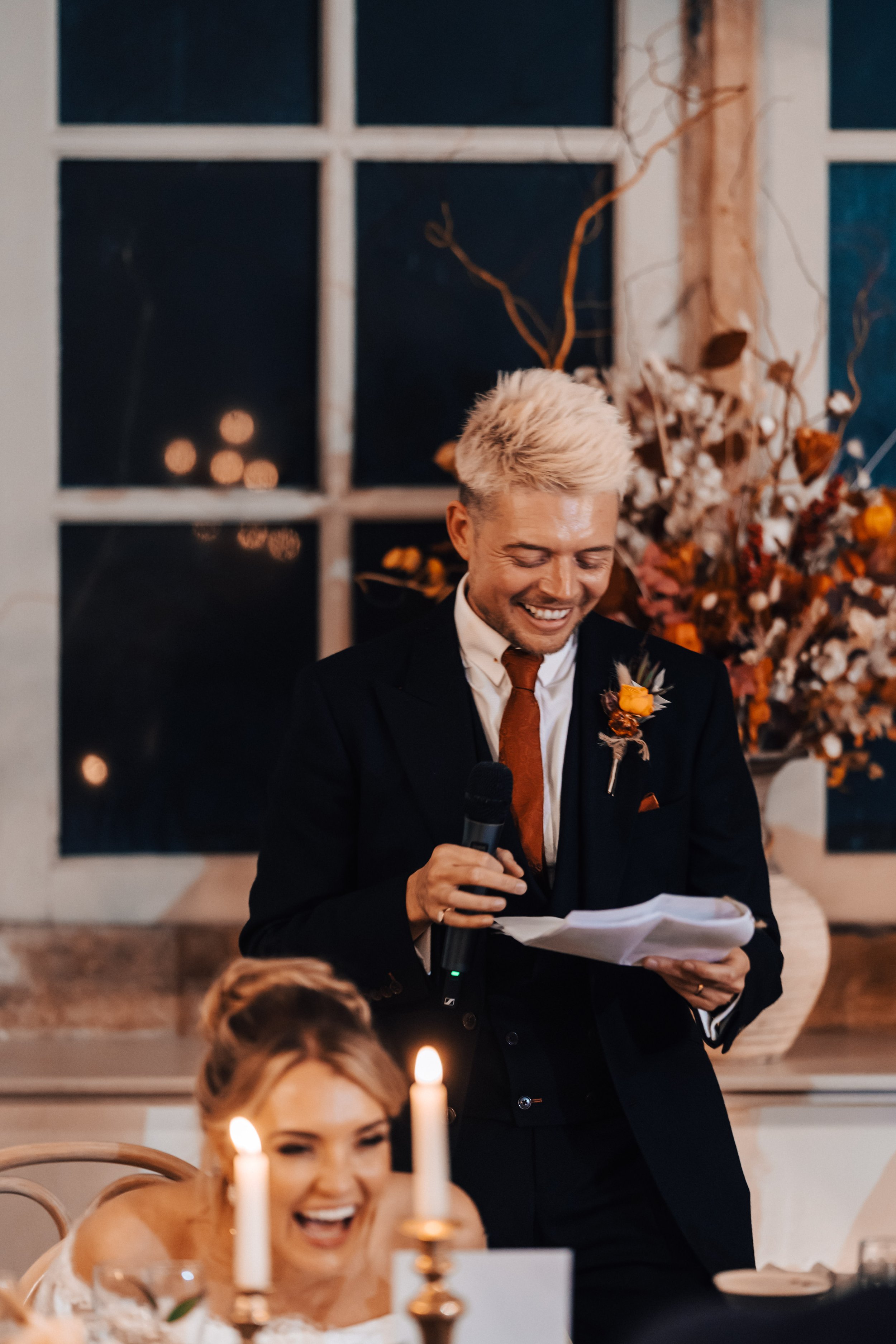 A man in a black suit and brown tie giving a speech with a microphone, smiling, during a wedding reception. A woman in wedding dress is in the foreground, laughing and seated at a table with lit candles.