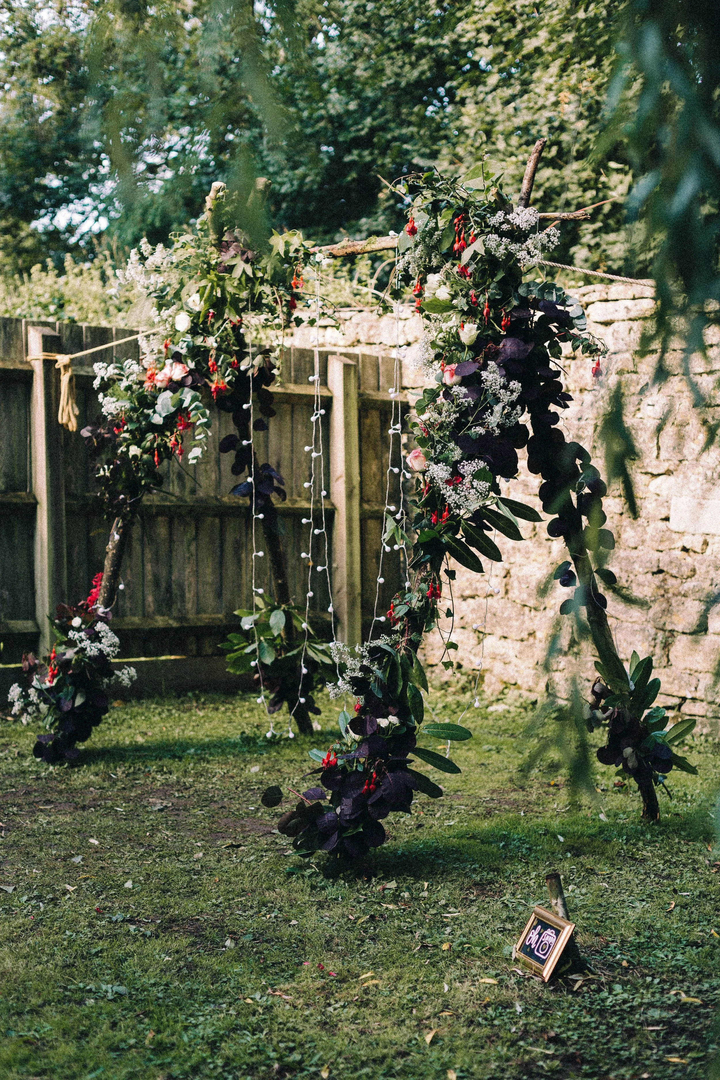 Decorated outdoor wedding arch with flowers, greenery, and string lights on a grassy lawn, with a wooden fence and stone wall in the background.