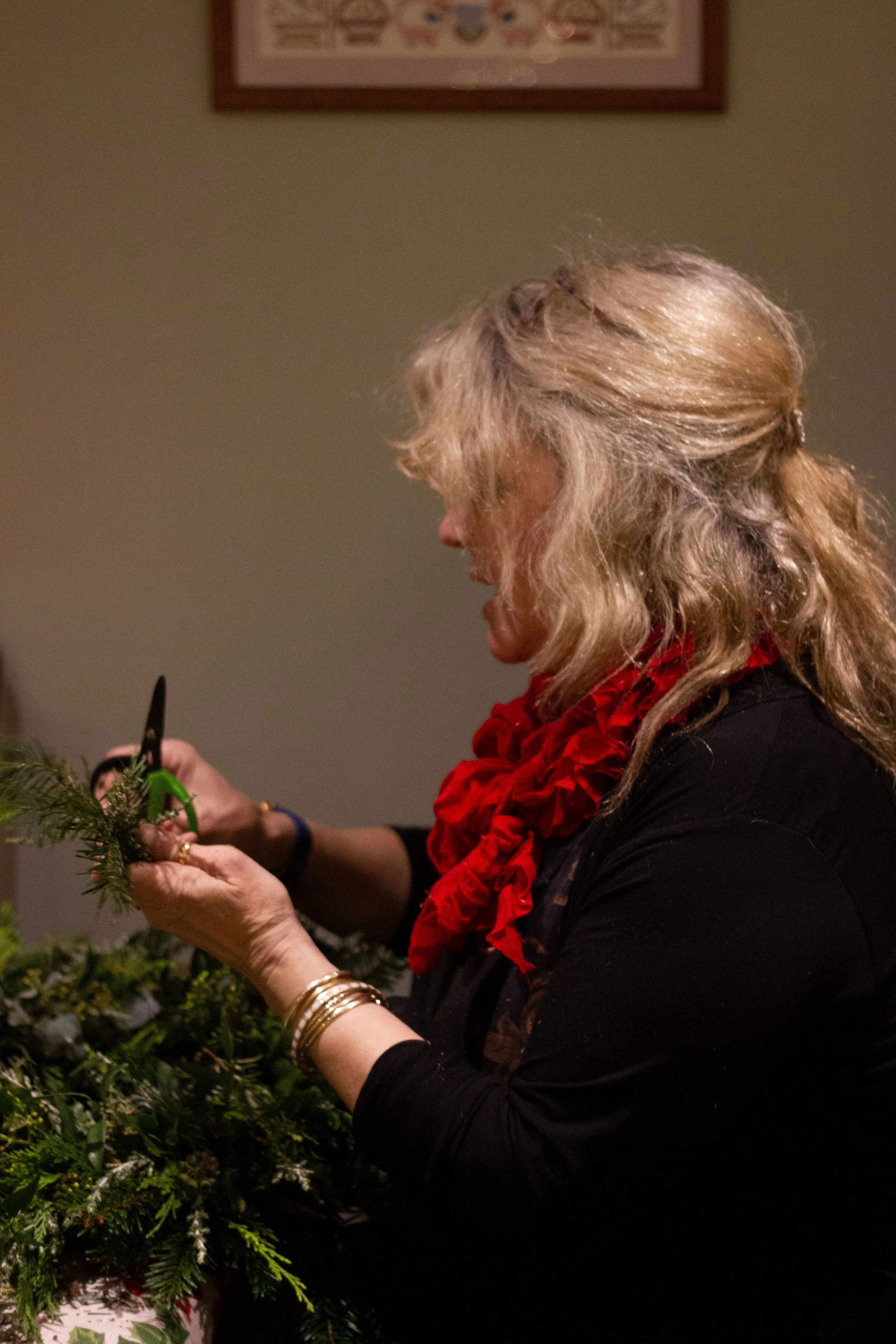 Woman with curly blonde hair, wearing a black top and red scarf, decorating or arranging holiday greenery.