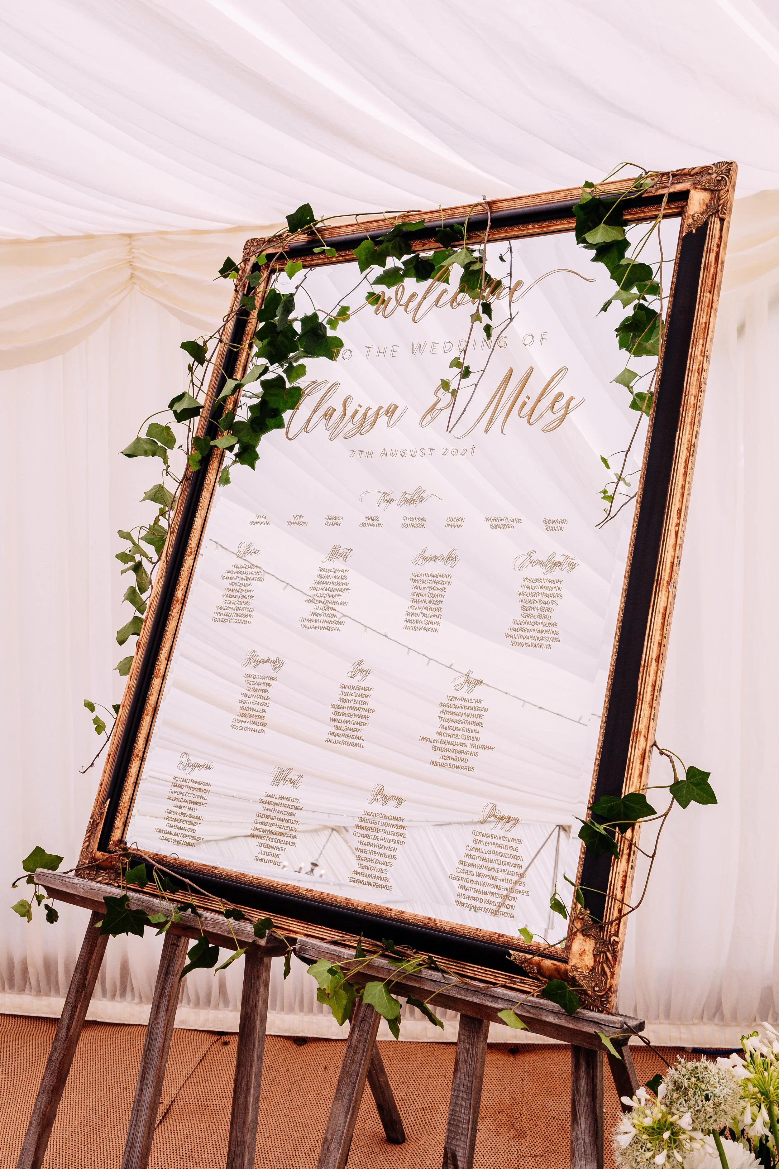 Wedding seating chart display on a large mirror with a wooden frame, decorated with green ivy, at a wedding reception.