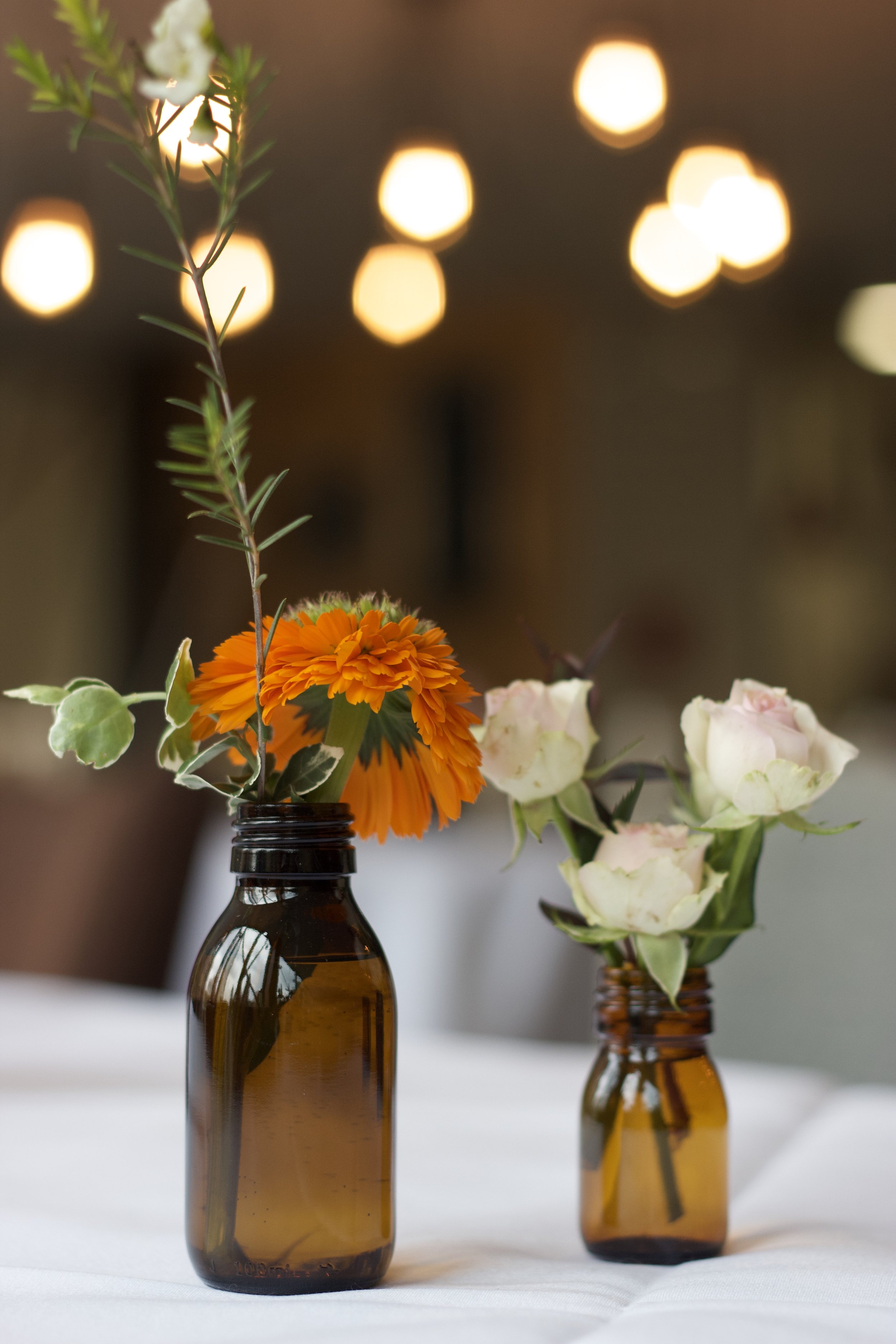 Two small amber glass bottles holding flowers, one with white roses and the other with orange and green flowers, on a white tablecloth with warm blurred lights in the background.