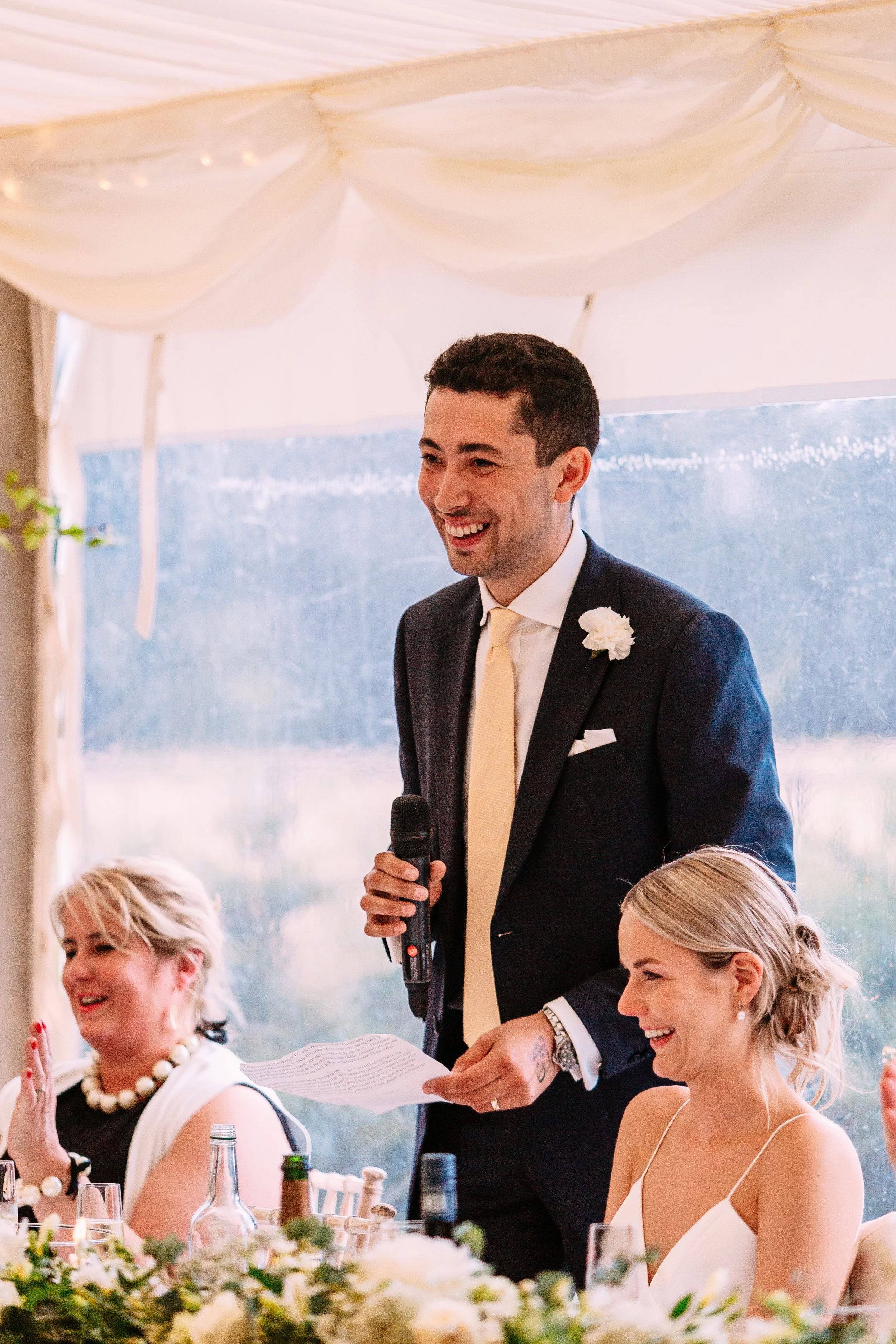 A man in a navy suit and yellow tie giving a speech at a wedding reception, holding a microphone and a piece of paper, with two women sitting at the table smiling and listening.