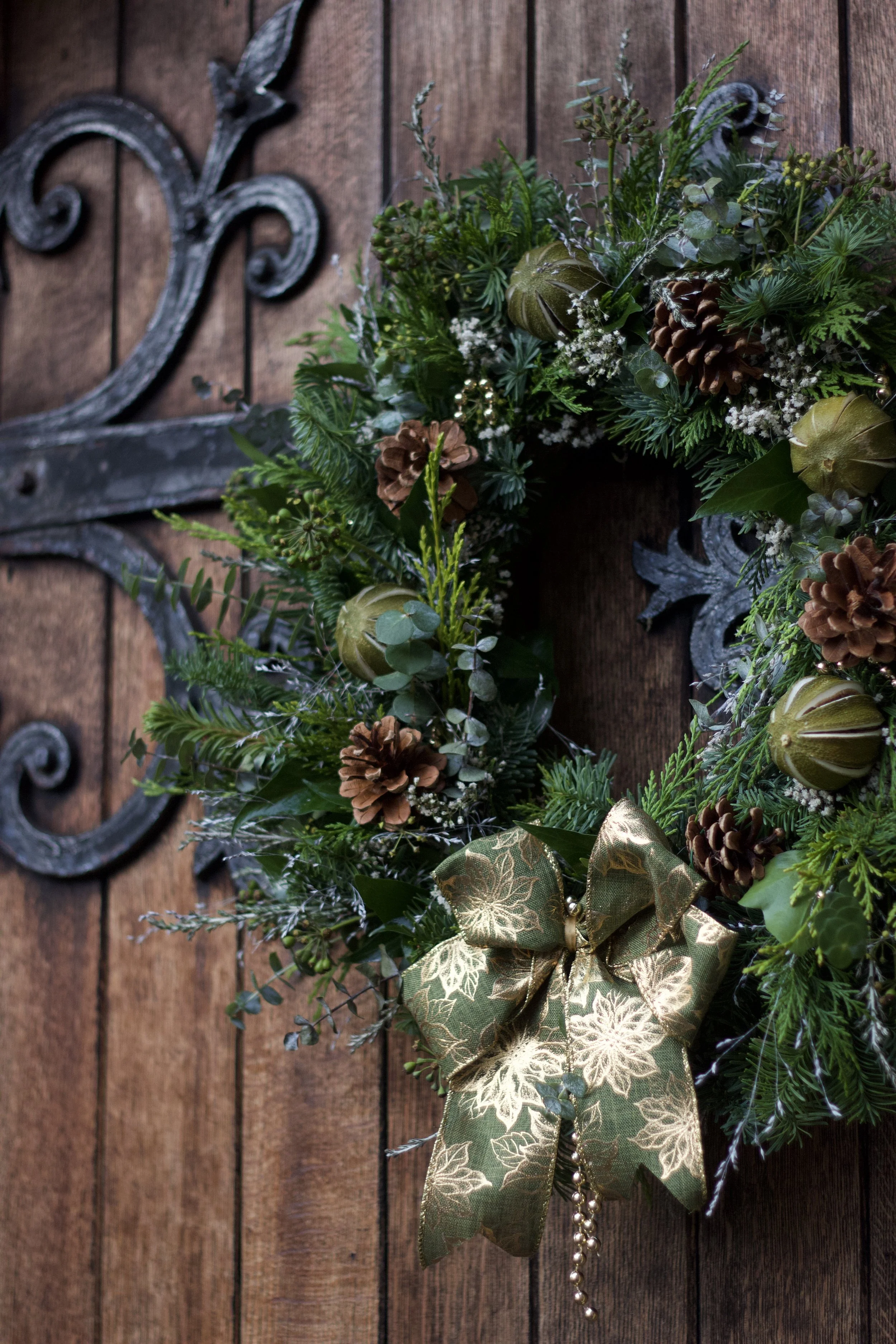 Christmas wreath with pinecones, green foliage, gold ribbon, and ornaments hanging on a wooden door.