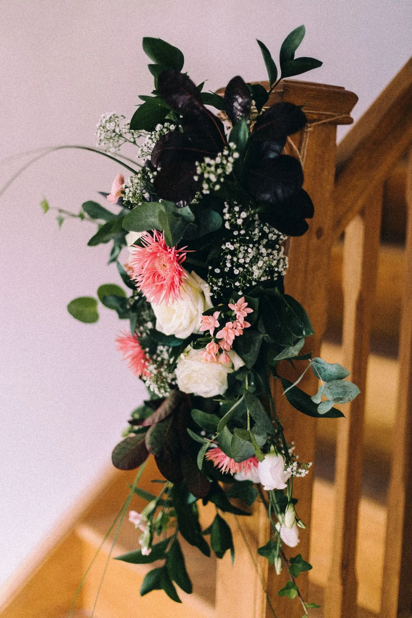 A floral arrangement with pink, white, and green flowers and dark green leaves, draped over a wooden stair railing.