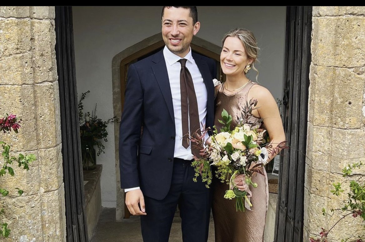 A happy couple standing in a doorway, dressed in formal attire, with the woman holding a bouquet of flowers, smiling and looking cheerful.
