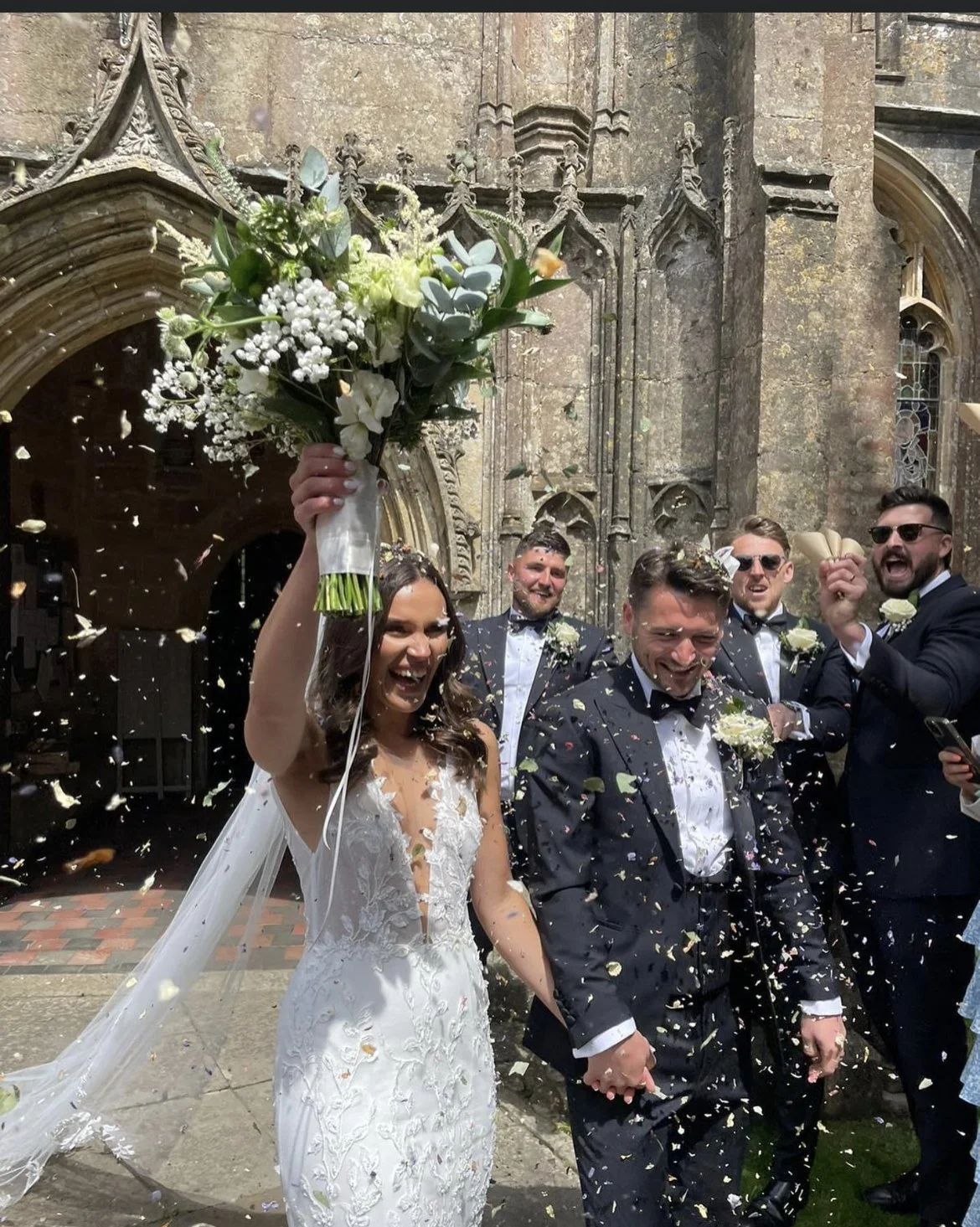 Bride and groom celebrating with friends outside a historic stone church, confetti in the air, bride holding a bouquet of white flowers, and everyone smiling.