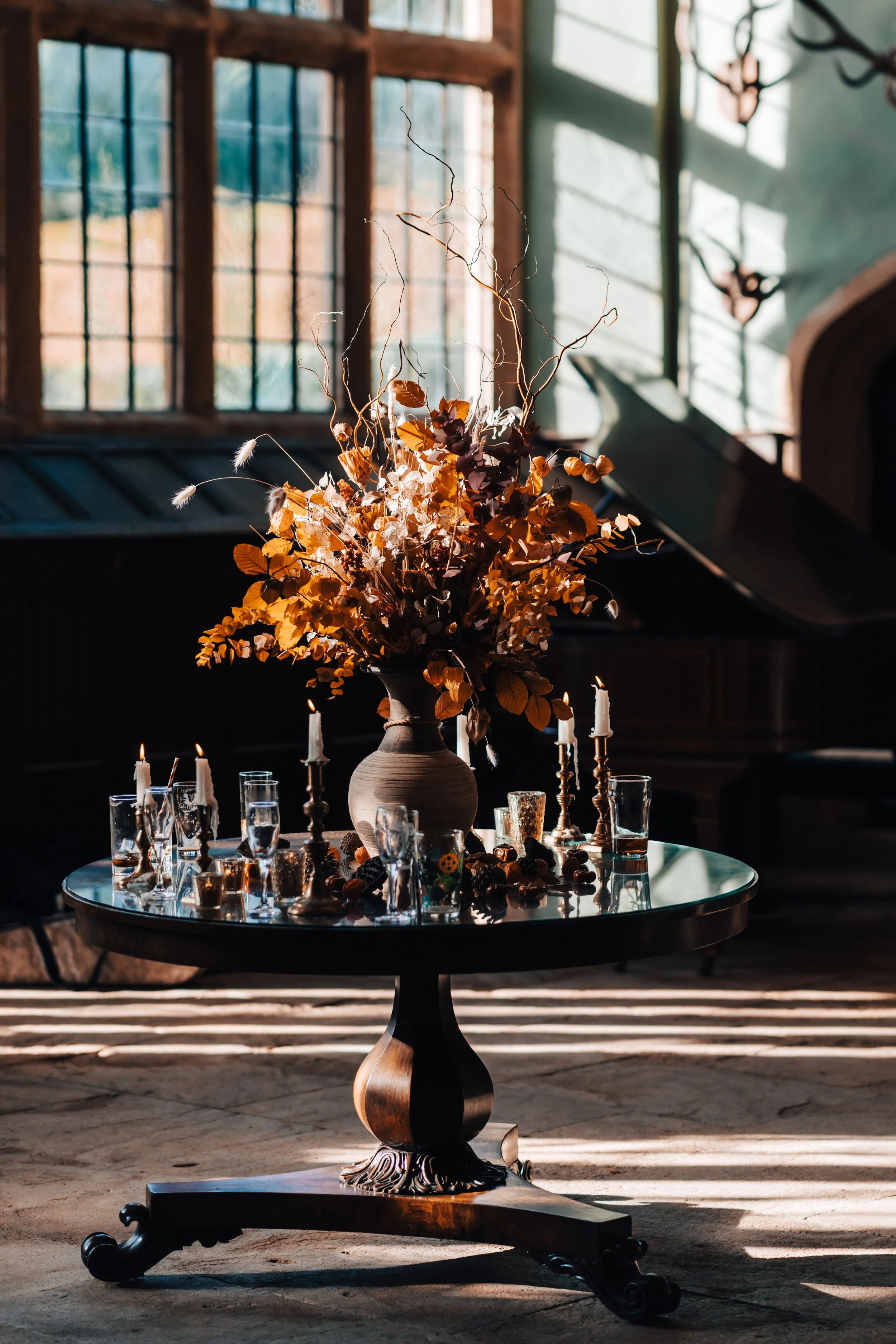A round wooden table with a large floral arrangement in a brown vase, surrounded by glass candles and drinking glasses, in a sunlit room with tall windows and dark wood paneling.