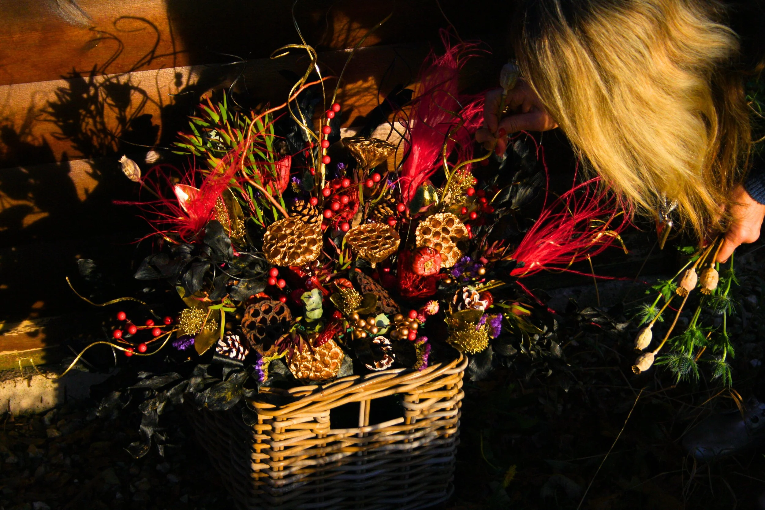 A woman with blonde hair arranging a floral and decorative arrangement in a basket, with golden, red, and purple accents, outdoors at sunset.