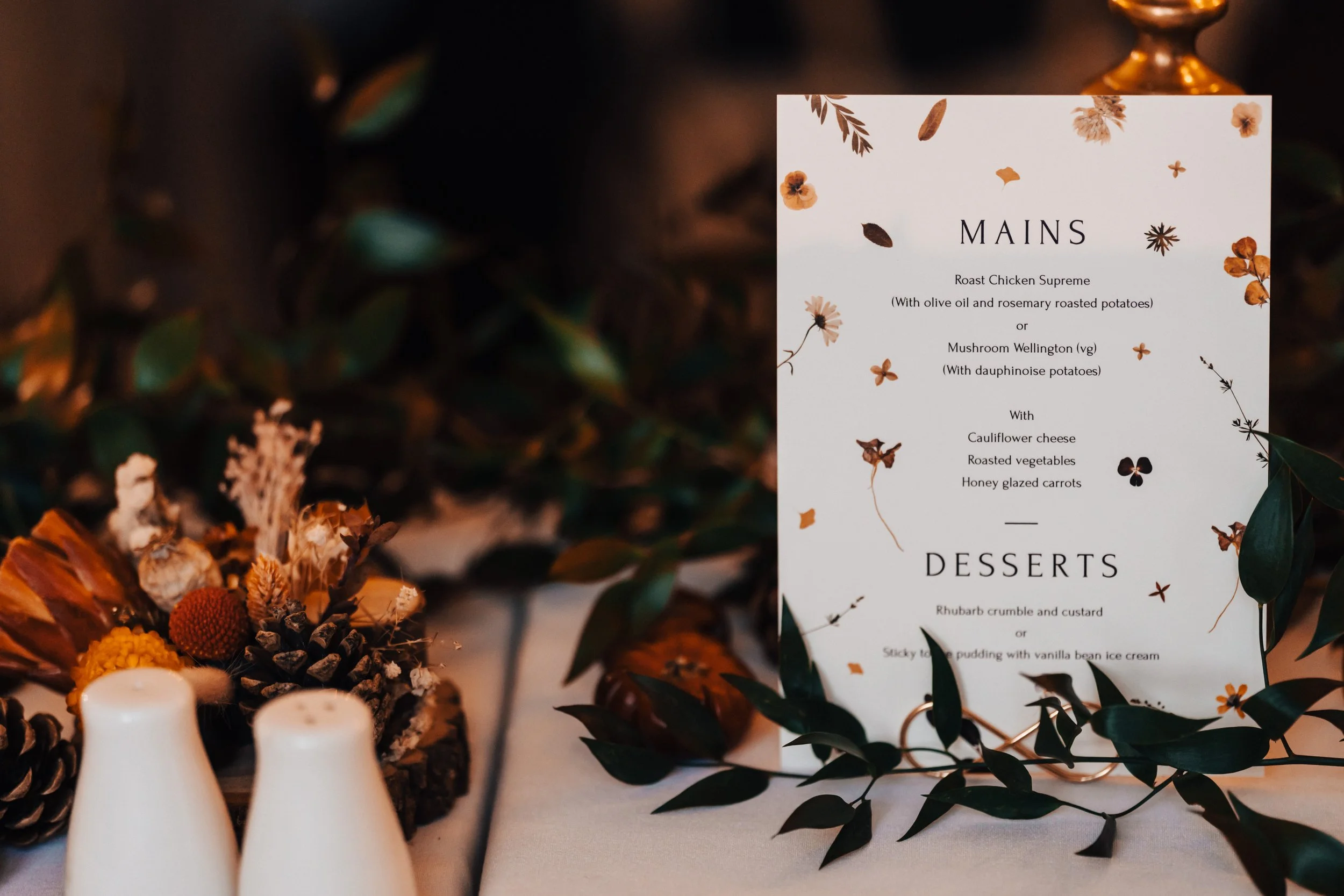 A menu card for a meal with floral and leaf decorations, positioned on a table with pinecones, dried flowers, and two white salt and pepper shakers.
