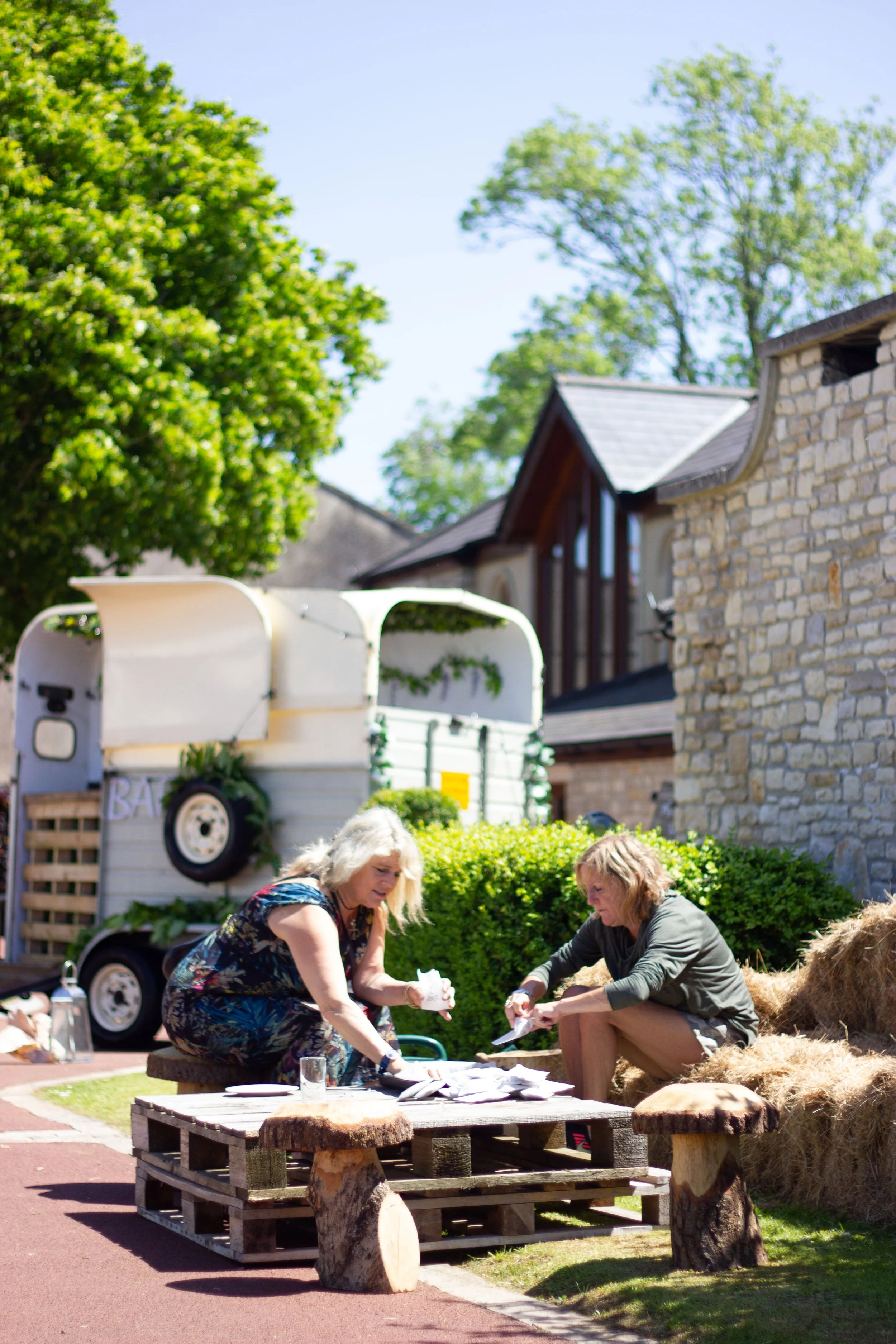Two women sitting outdoors at a picnic table, engaging with papers or crafts, with a vintage trailer decorated with greenery and potted plants in the background, surrounded by trees and a stone building.