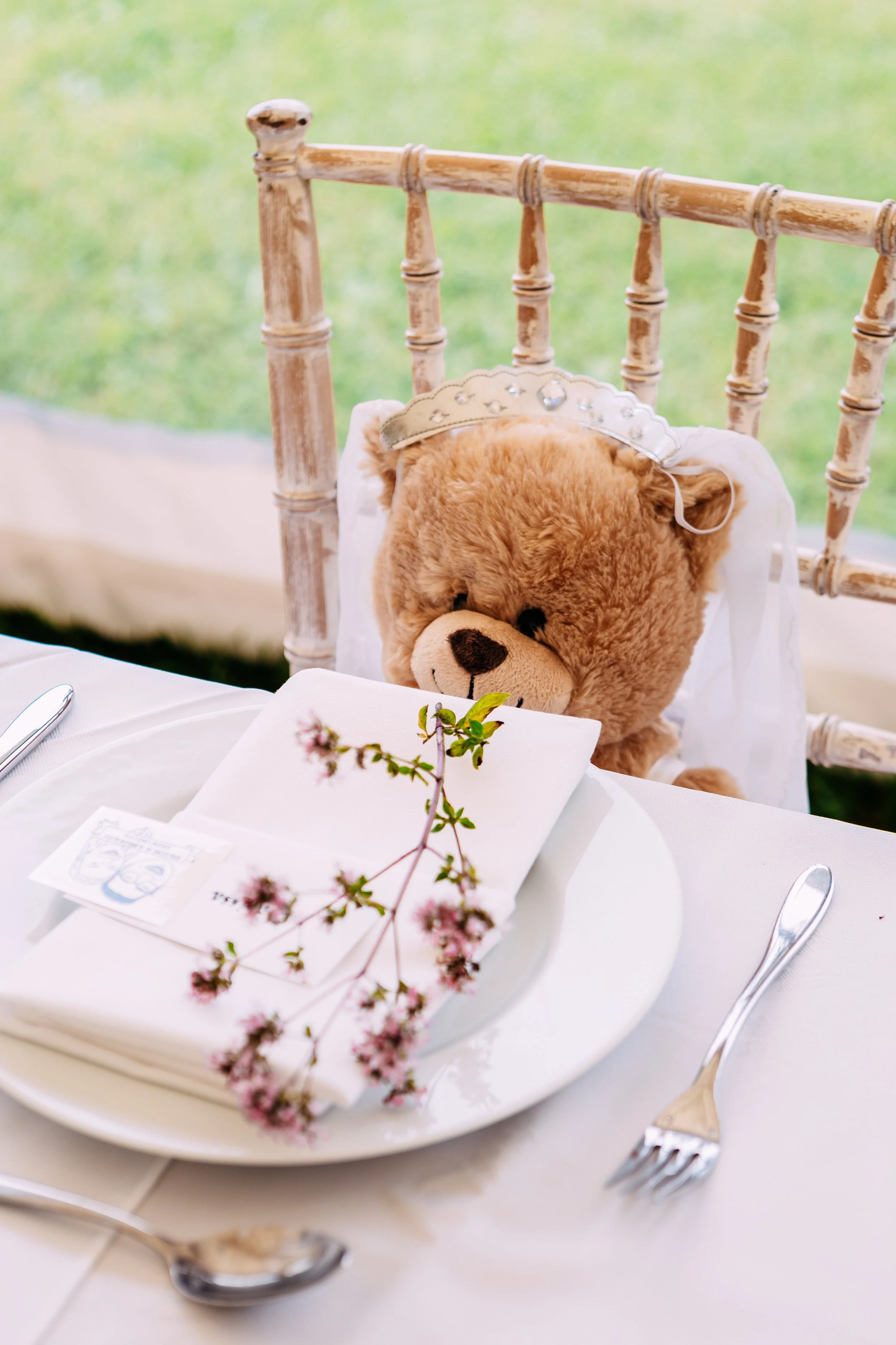 A teddy bear wearing a tiara sits at a dining table with a white tablecloth, set with a plate, napkin, and cutlery, against a blurred outdoor background.