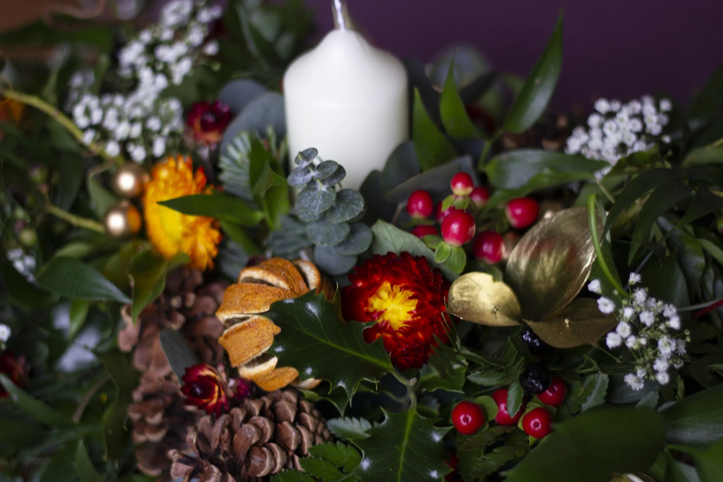 A festive Christmas arrangement with a white candle surrounded by holly leaves, red berries, pinecones, gold and red decorative elements, and small white flowers.