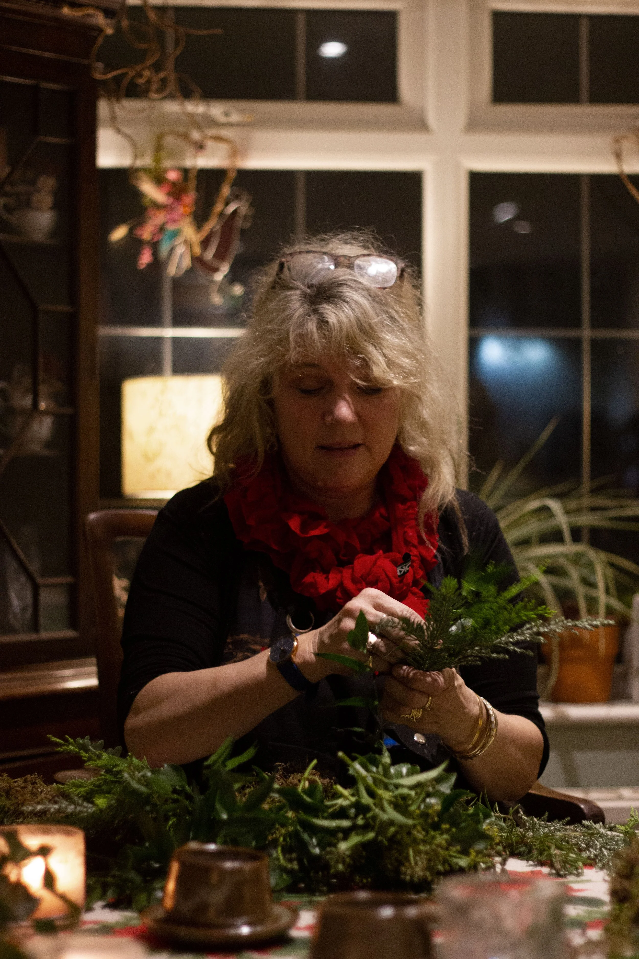 A woman with blonde, curly hair and glasses on her head, wearing a black top with a red ruffled scarf, sitting at a table arranging green pine branches and foliage for a holiday centerpiece in a warmly lit room.