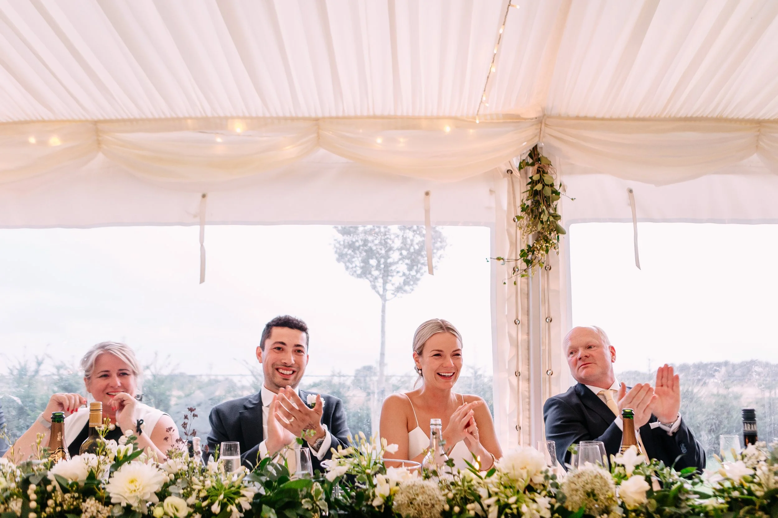 People sitting at a decorated table during a celebration, possibly a wedding reception, inside a white tent with hanging curtains and greenery.