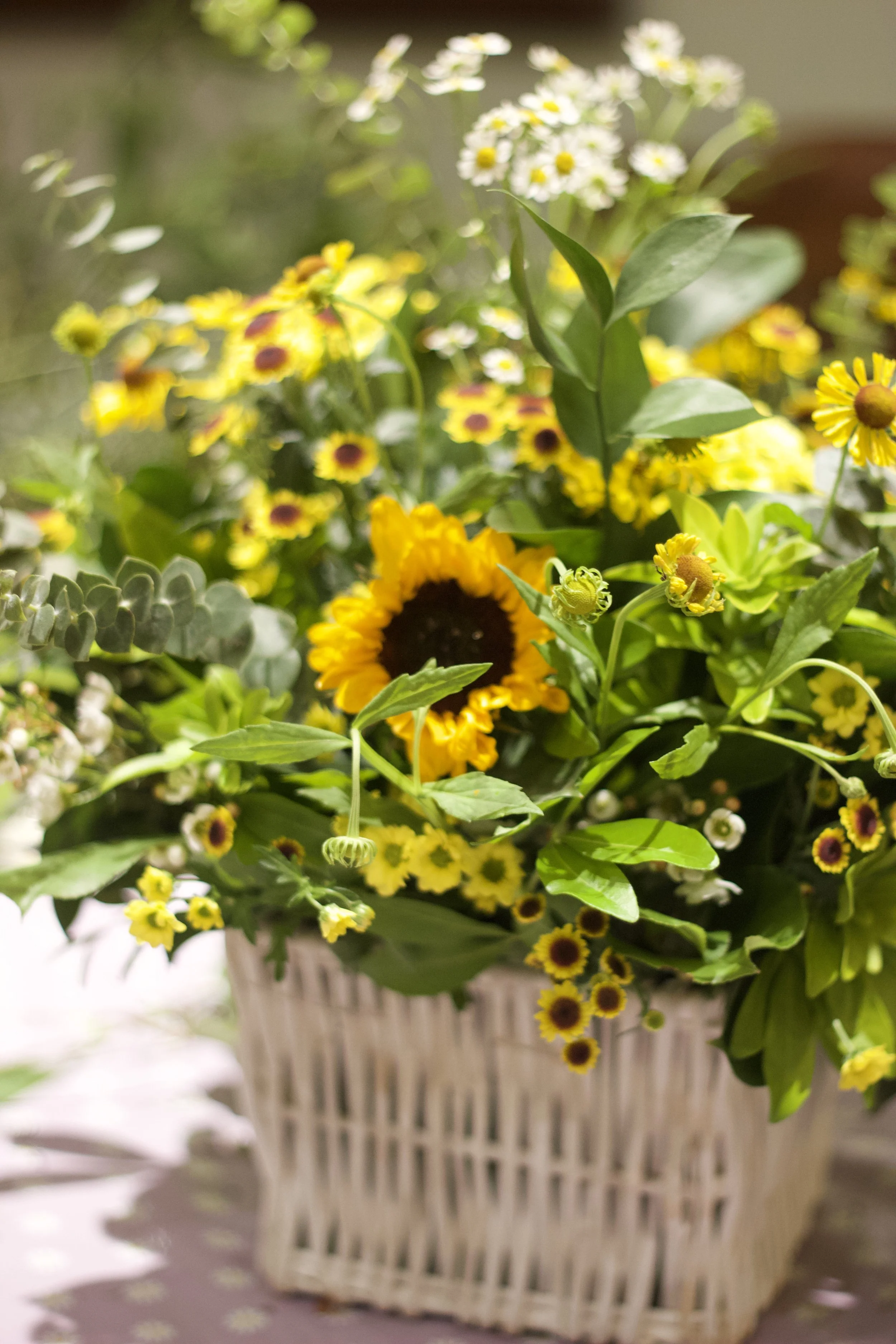 A basket filled with a vibrant flower arrangement including sunflowers, small white and yellow flowers, and greenery.