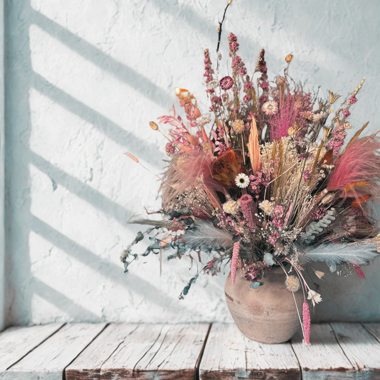 Dried floral arrangement in a round, beige clay vase on a rustic wooden surface against a textured white wall, with sunlight and shadows.