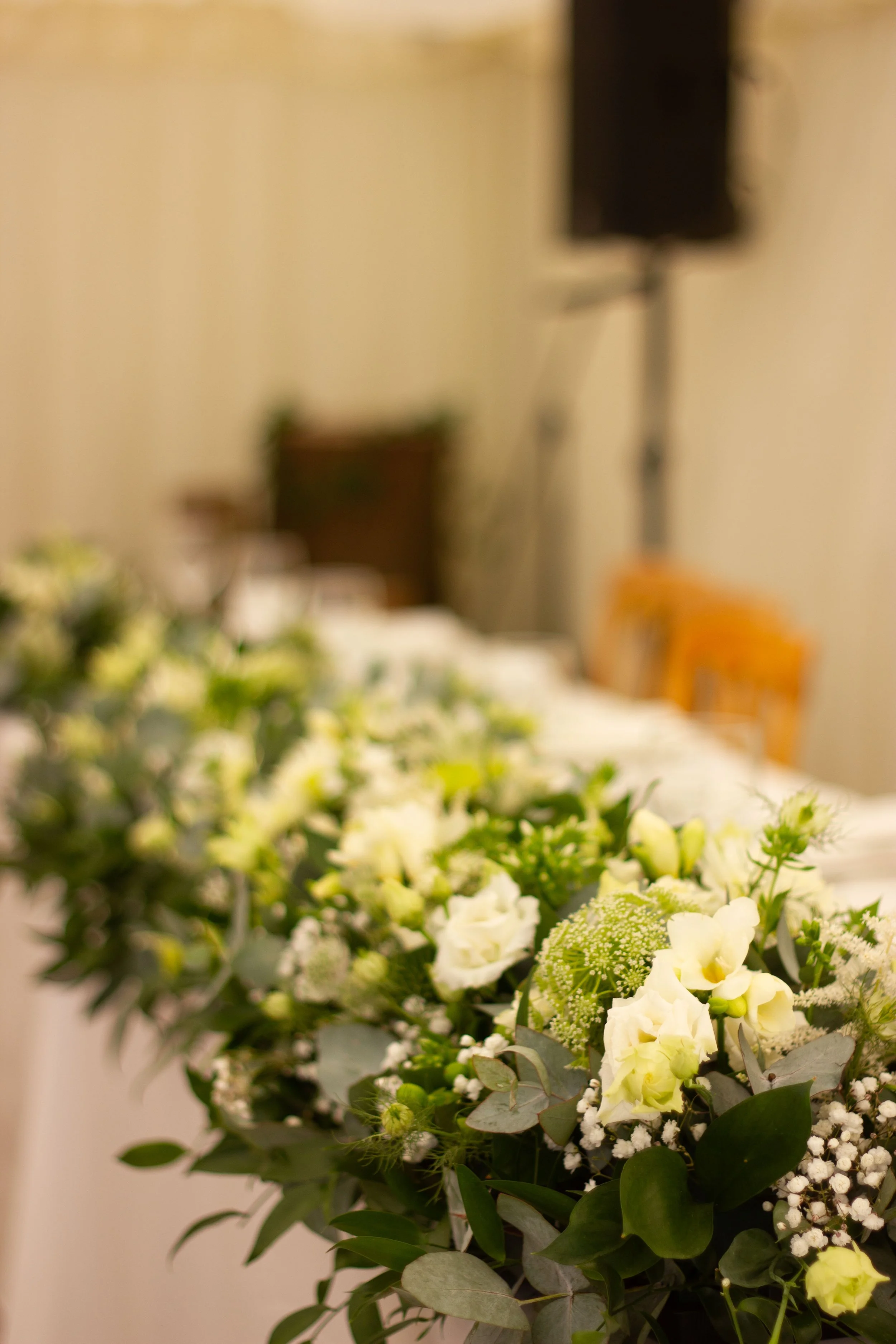 A floral arrangement of white flowers and greenery on a table in an indoor setting, with a blurred background.