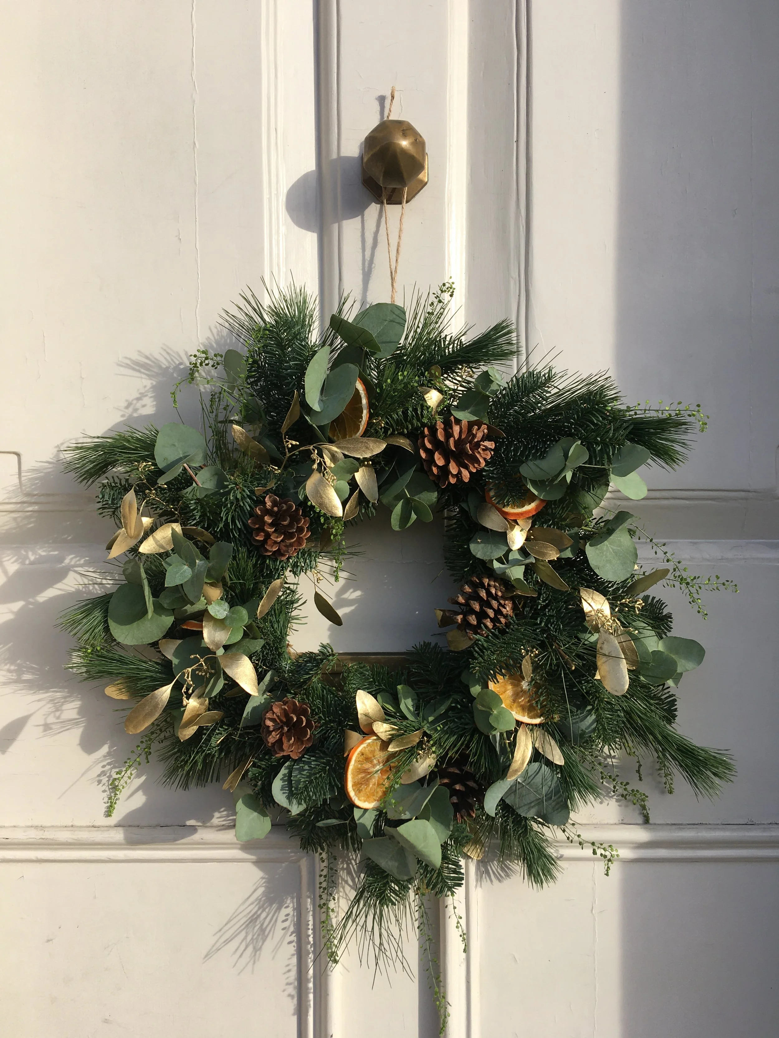 A holiday wreath with greenery, pinecones, dried orange slices, and gold-painted leaves hanging on a white door.