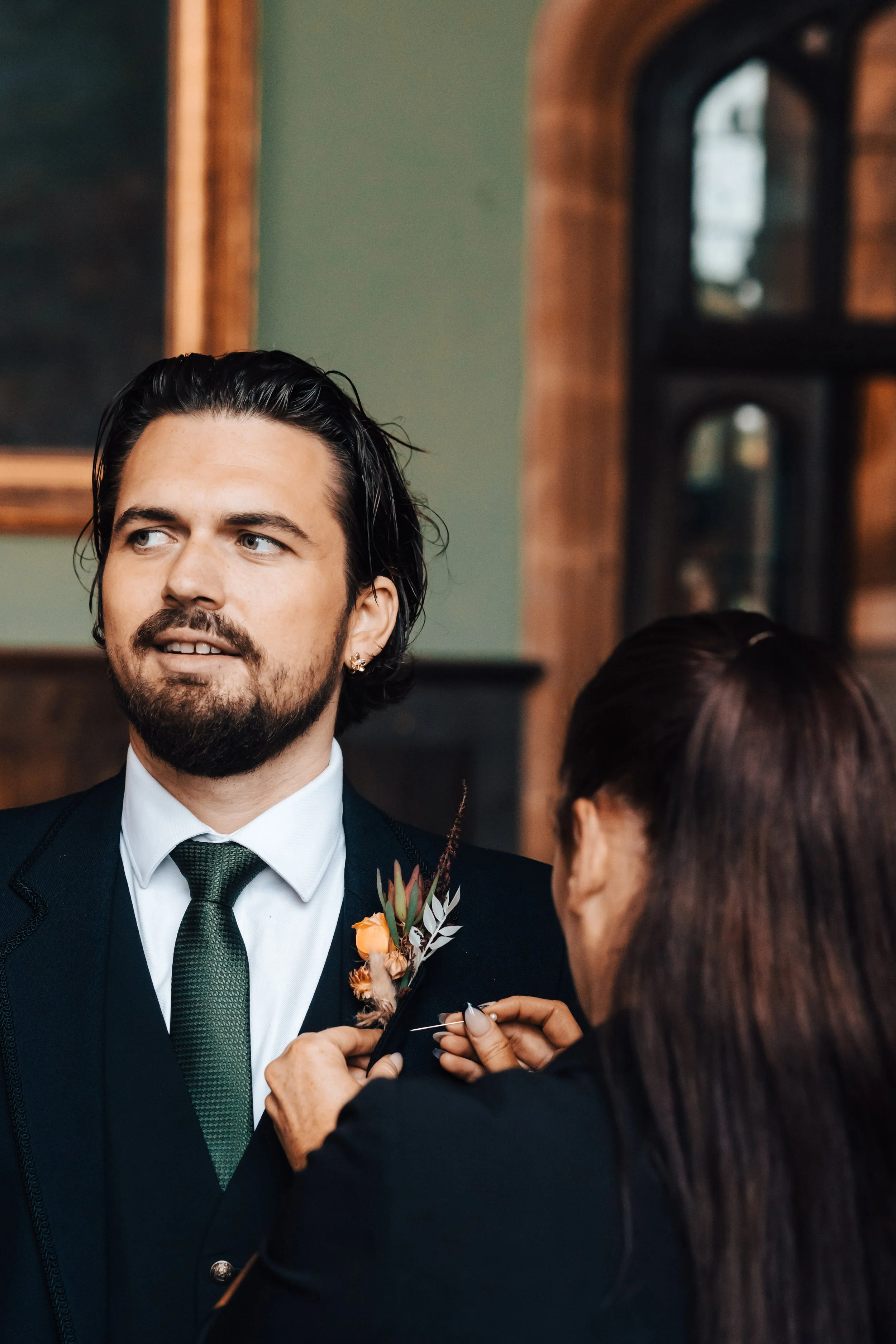 A man in a formal black suit with a white shirt and green tie getting his boutonniere pinned on during a wedding ceremony, with a woman assisting him.