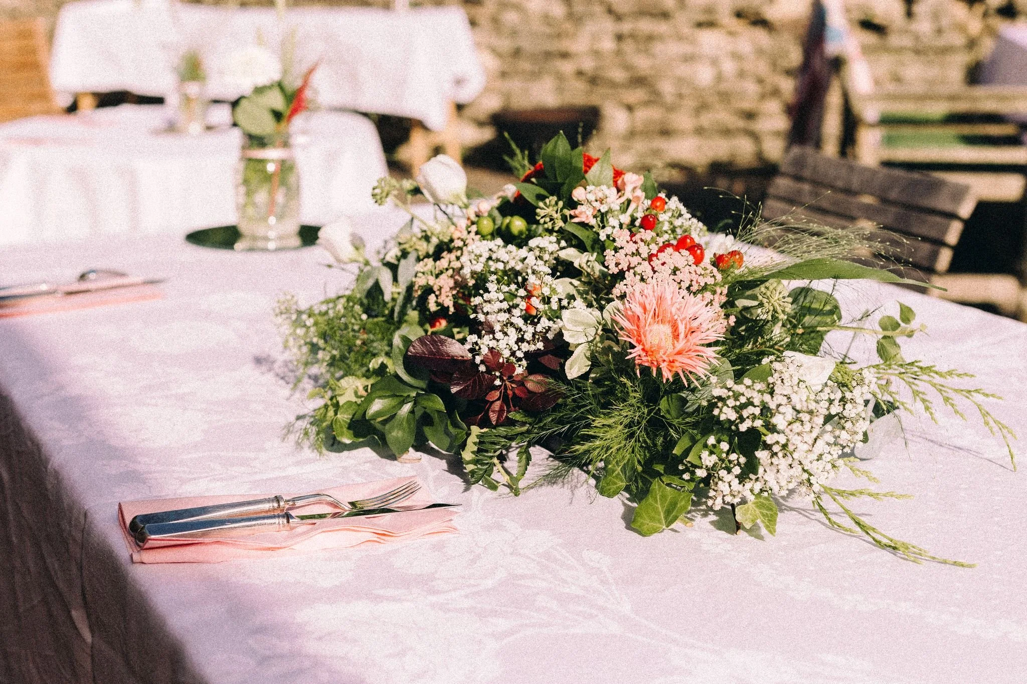 A table with a pink floral centerpiece surrounded by greenery and small flowers, set for a meal with utensils wrapped in pink napkins.