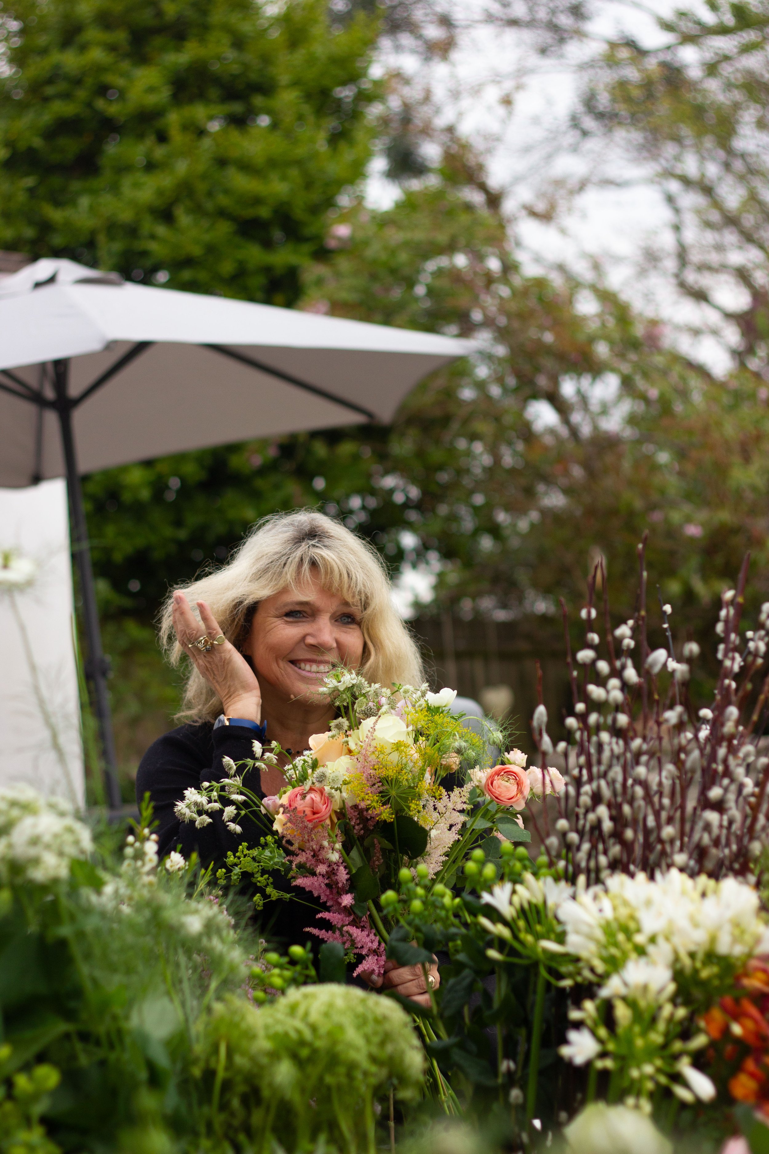 A woman with blonde hair smiling and holding a bouquet of flowers outdoors in a garden, with trees and a white patio umbrella in the background.