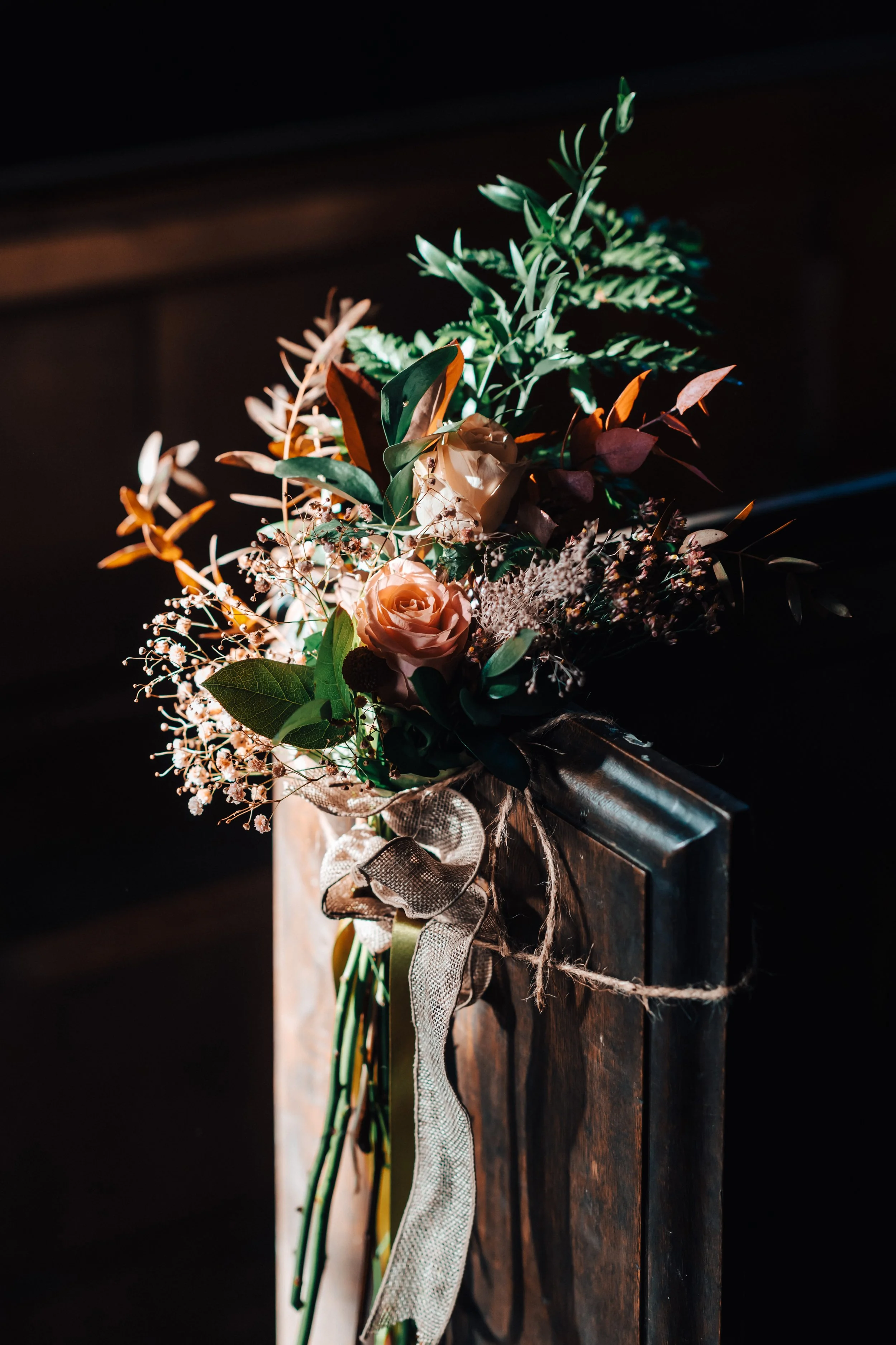 A bouquet of pink roses and assorted greenery with ribbon tied to a wooden surface.