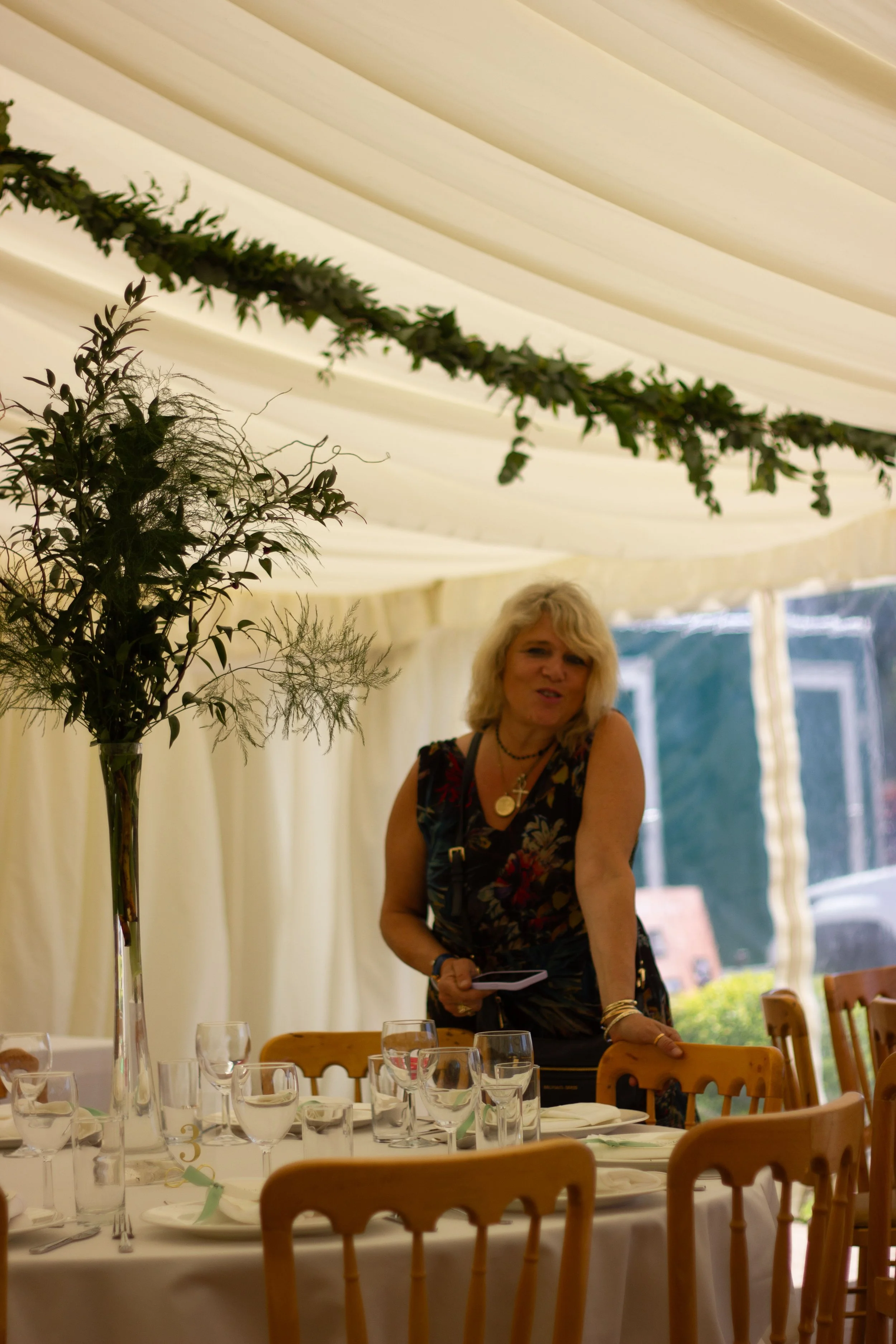 Woman in a sleeveless floral dress standing next to a dining table inside a decorated tent, with glasses and a tall floral centerpiece on the table, and greenery draped from the ceiling.
