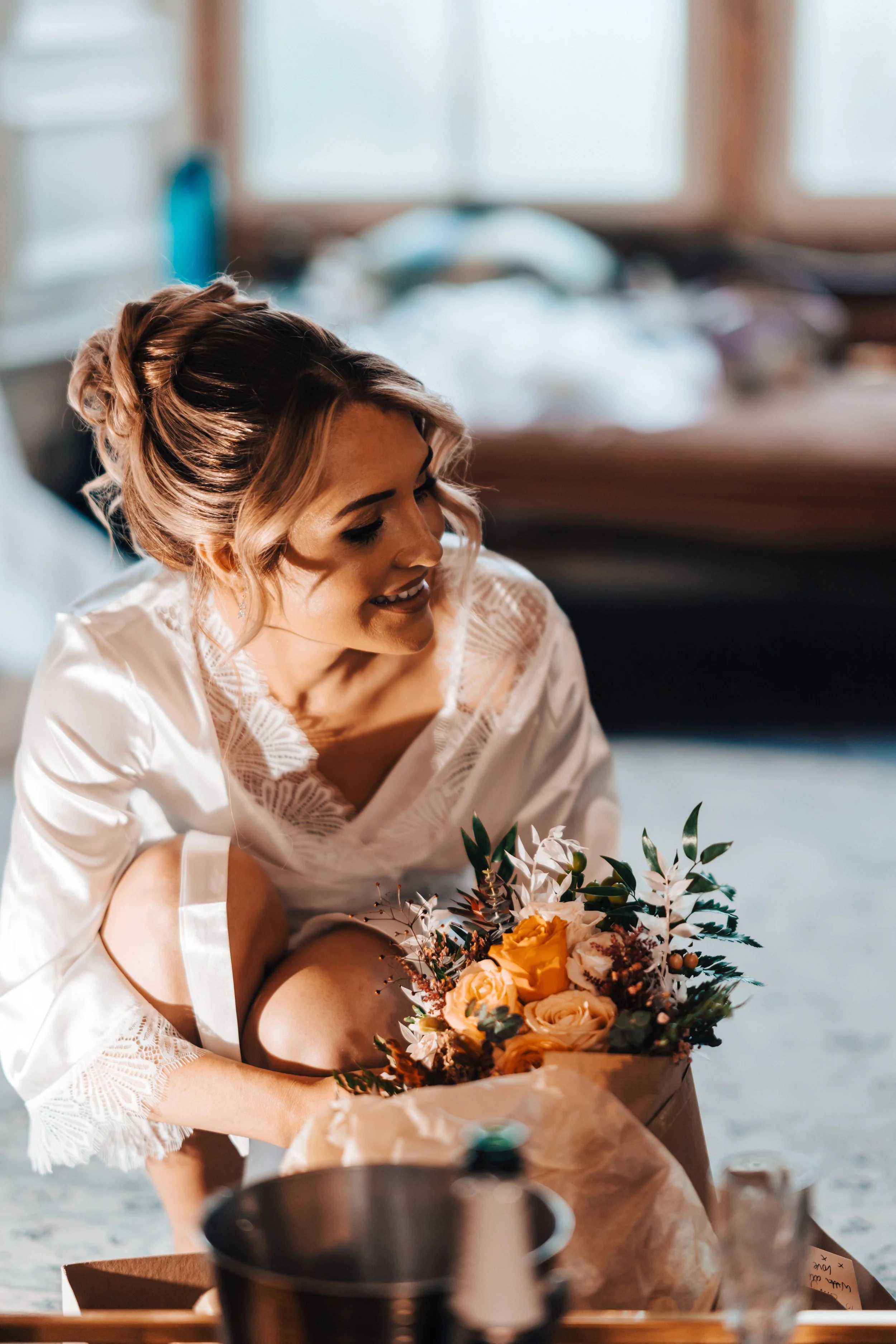 A woman with styled hair and a white lace robe sitting on the floor, holding a bouquet of flowers, in a cozy indoor setting with a blurred bed in the background.