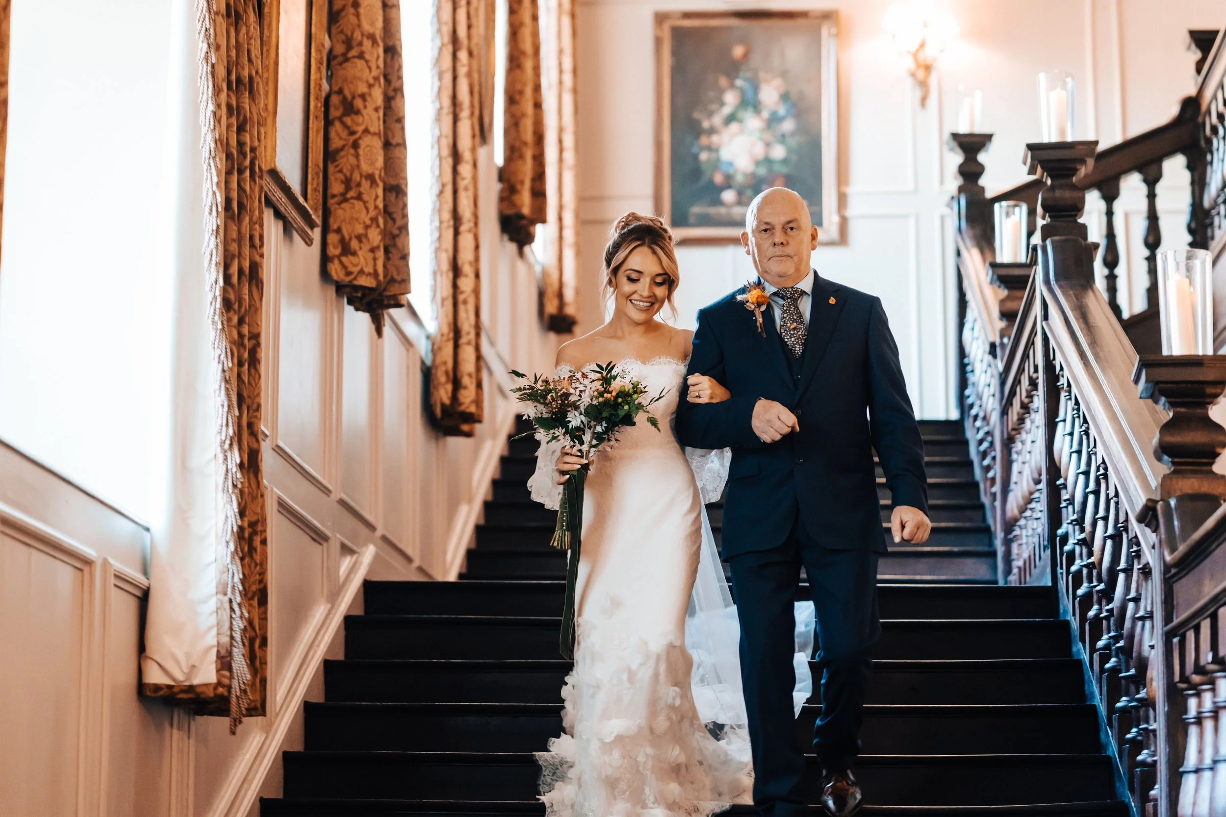 A bride smiling and holding a bouquet, walking down the stairs with an older man in a suit, in a decorated indoor setting with ornate curtains and paintings.