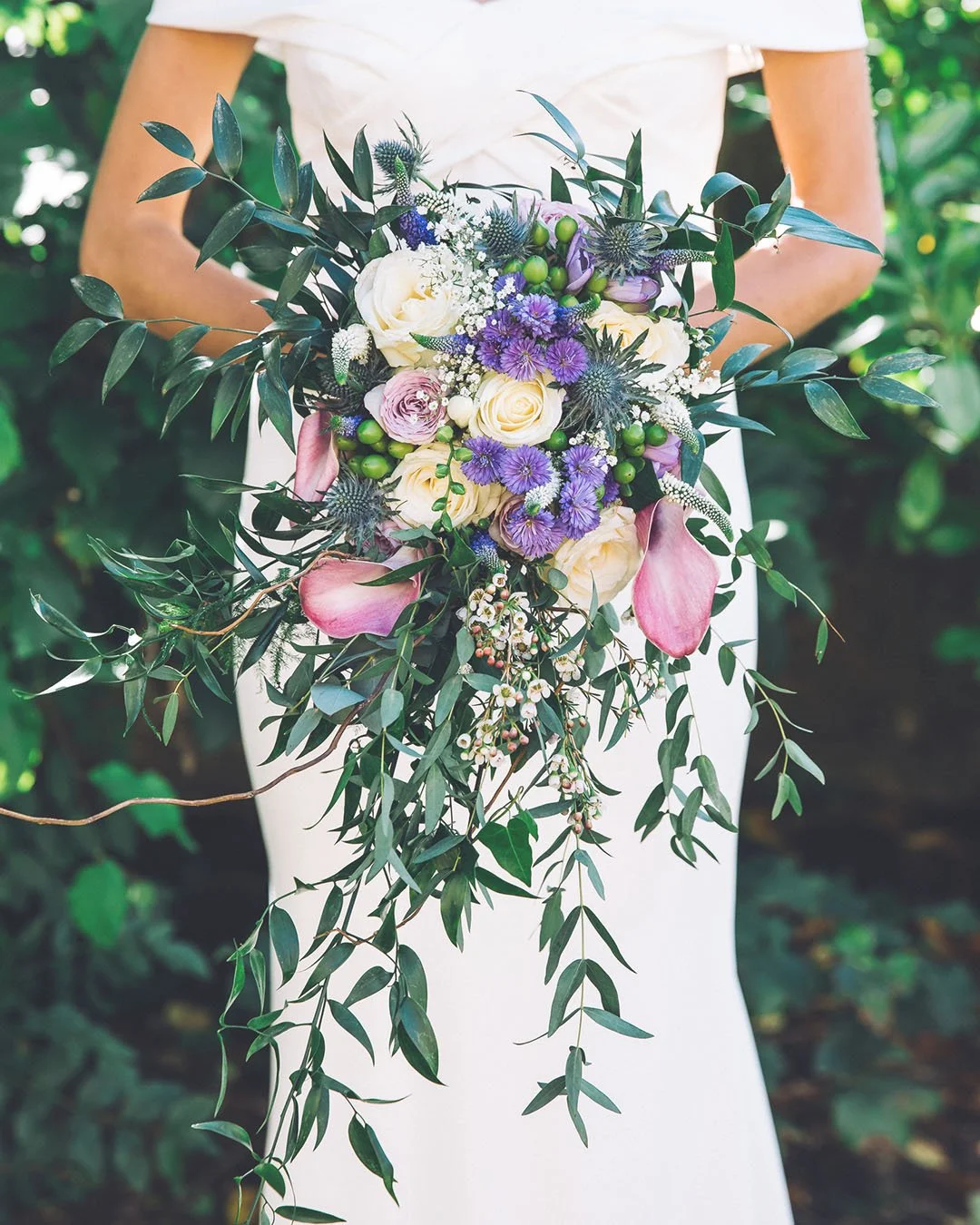 A person in a white dress holding a cascading bridal bouquet with white roses, purple flowers, pink calla lilies, greenery, and small white filler flowers.
