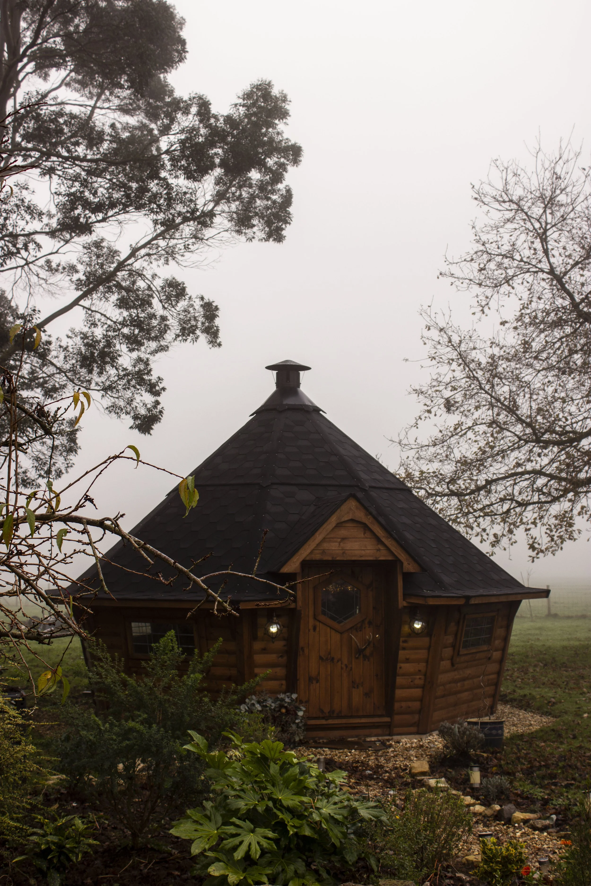 A small, round wooden house with a conical black roof and a chimney, surrounded by trees and plants, on a foggy day.