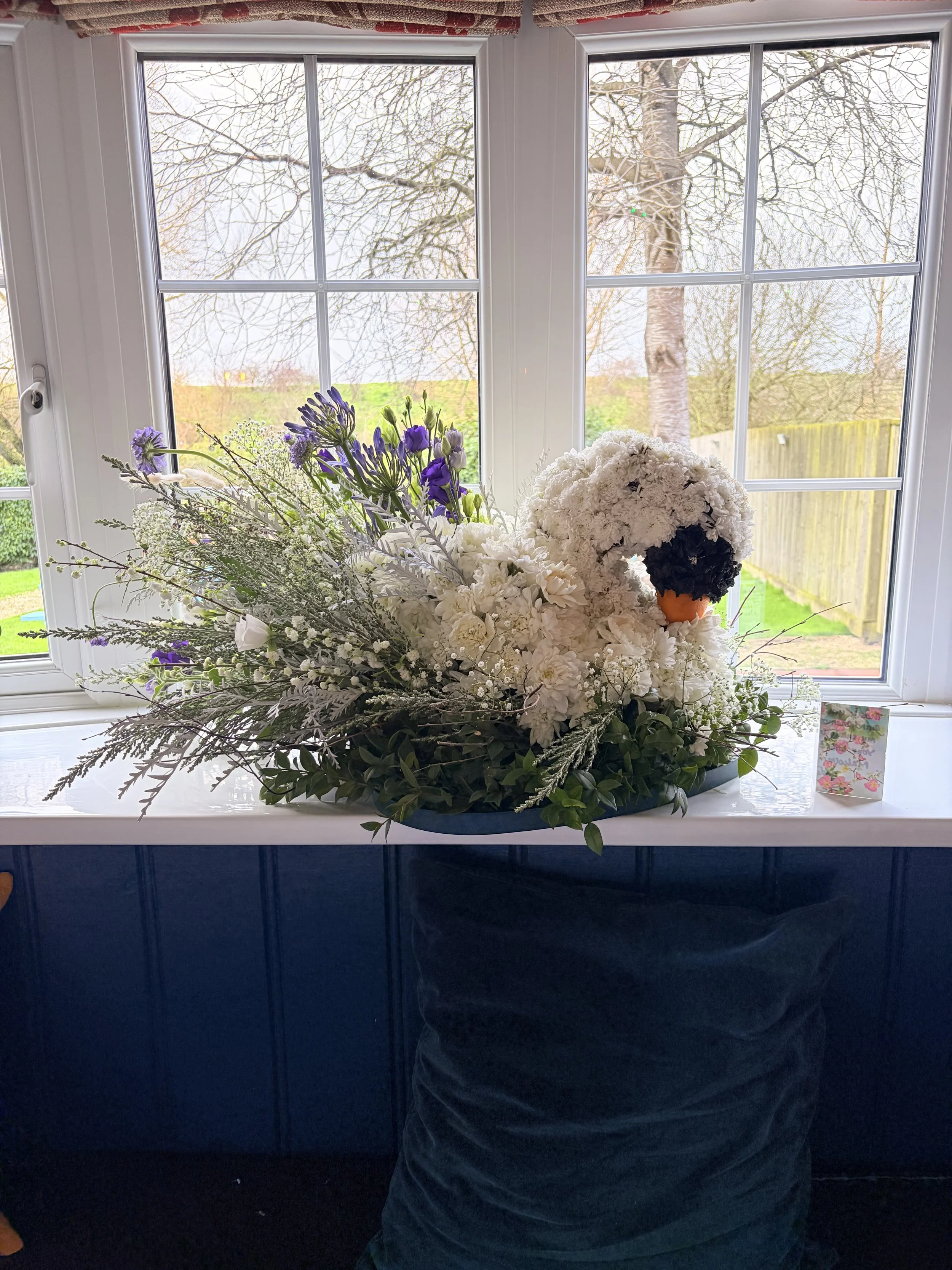A floral arrangement on a windowsill featuring a swan made of white flowers with an orange beak and black eyes, surrounded by various greenery and purple flowers, with a window revealing a backyard with trees outside.